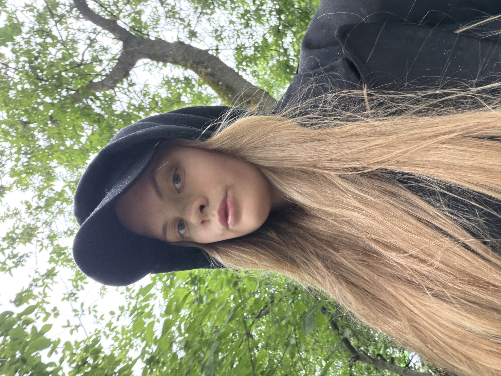 An outdoor portrait features a young person, likely female, captured from a low angle looking upwards. The subject is centered in the frame, with their head tilted significantly, making the image appear rotated. They have long, straight, light blonde or light brown hair that cascades down their shoulder and chest, appearing slightly damp or dusty with fine white specks. The person is wearing a black hooded garment, possibly a jacket or sweatshirt, with the hood up, framing their face. Their eyes are light-colored, and a neutral or calm expression is visible on the partially obscured face, showing the nose and lips.
The setting is outdoors, beneath a tree with a visible trunk and abundant green foliage, suggesting a natural environment such as a park or garden in Izhevsk, Russia. The background is a bright, overcast sky seen through the tree canopy, providing soft, diffused lighting. The leaves are vibrant green, indicating a spring or summer season. Fine white specks are also visible on the black hooded fabric, consistent with the specks on the hair, which could be raindrops, pollen, or dust, hinting at the weather conditions during the shoot. No text is visible in the image.