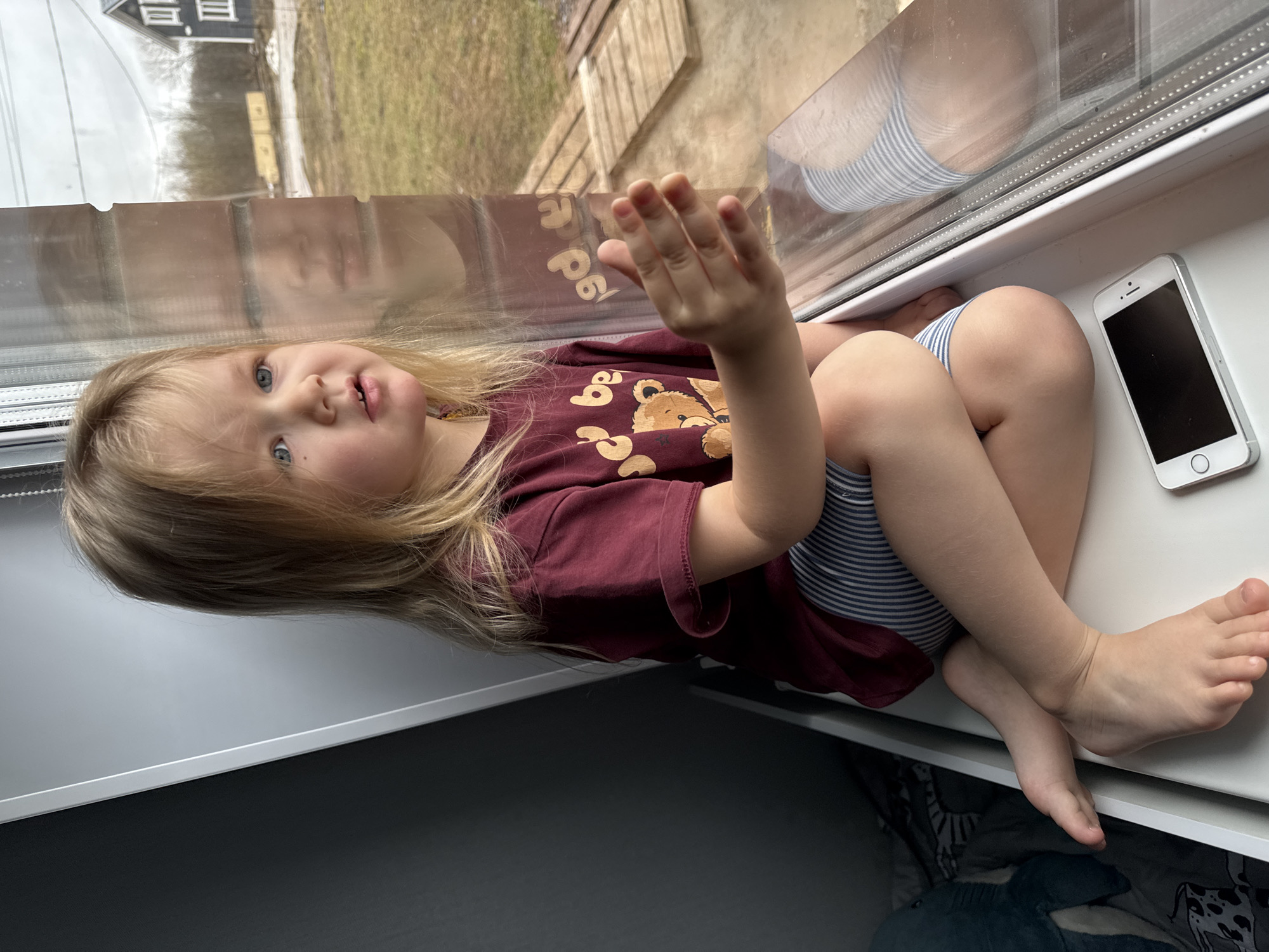 A young child with blonde hair and blue eyes sits on a white windowsill, looking towards the camera. The child wears a maroon t-shirt with a teddy bear graphic and striped shorts. The child's right hand is raised towards the window. The reflection in the window shows the child's face and parts of the outside: a house, and possibly a yard with some greenery. A white smartphone rests on the windowsill, beside the child. The windowsill appears to be inside a room with a gray surface underneath. Based on the reflection in the window, the location might be Gornyi (Dvurechenskoe S/P), Russia.