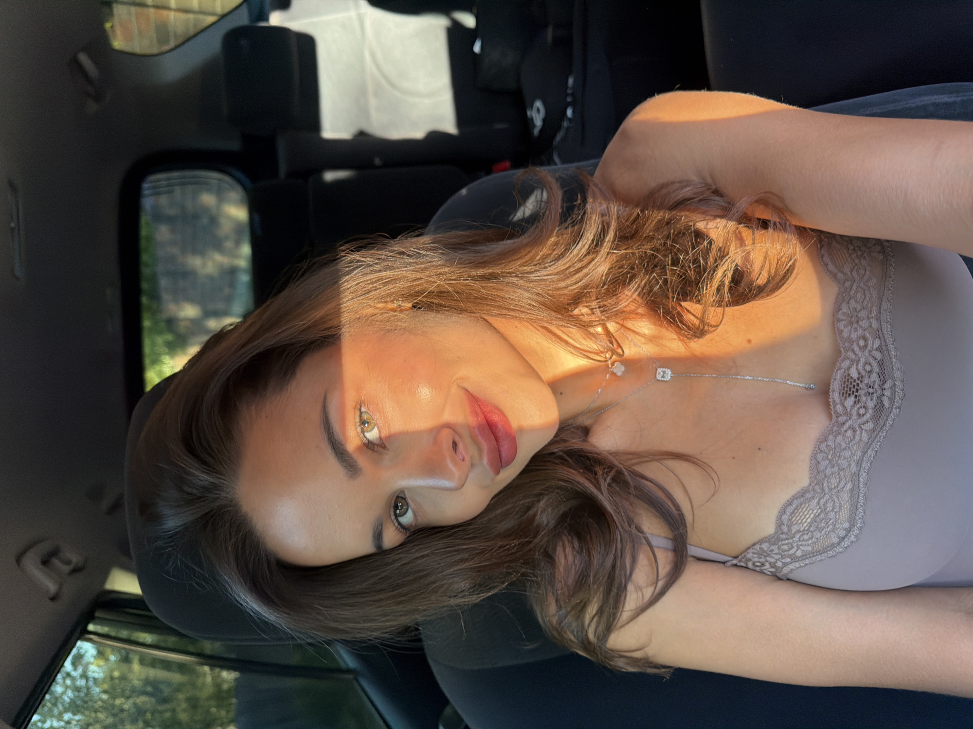 A young woman with light brown hair and fair skin is lying on her left side in a car. She has green eyes and red lips. She is wearing a light-colored top with lace trim and a silver necklace. The car is parked, and the background shows greenery, suggesting it might be a residential area or a park. The photo does not provide sufficient information to confirm the location is Krasnoyarsk, Russia.