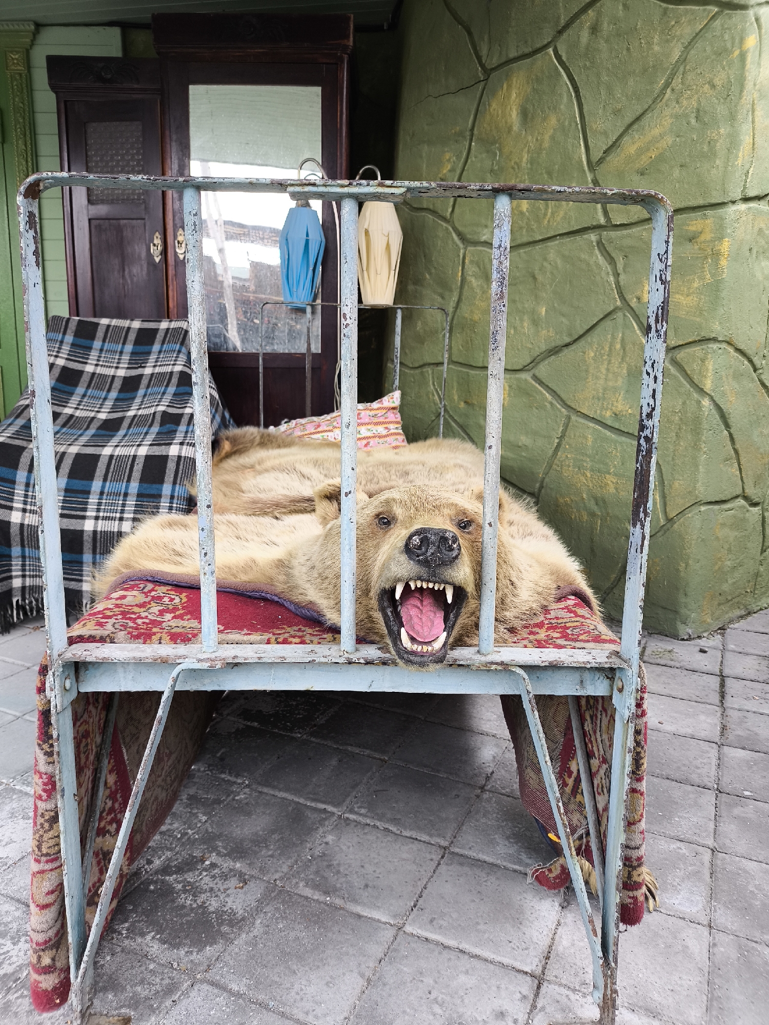 A taxidermied bear is positioned on a rusty, light blue metal bed frame. The bear is lying on its back, with its mouth open, revealing sharp teeth and a pink tongue. It is covered by a red rug with a geometric pattern. Another rug, a blue and black plaid blanket, is draped over the head of the bed frame behind the bear. In the background, there is a dark wooden cabinet with a mirrored door, and a textured green wall that resembles rock formations. The scene is set outdoors, on a paved area, with the overall impression being that of a roadside attraction or a display in a rural setting. The location is specified as Borgustanskaya, Russia.
