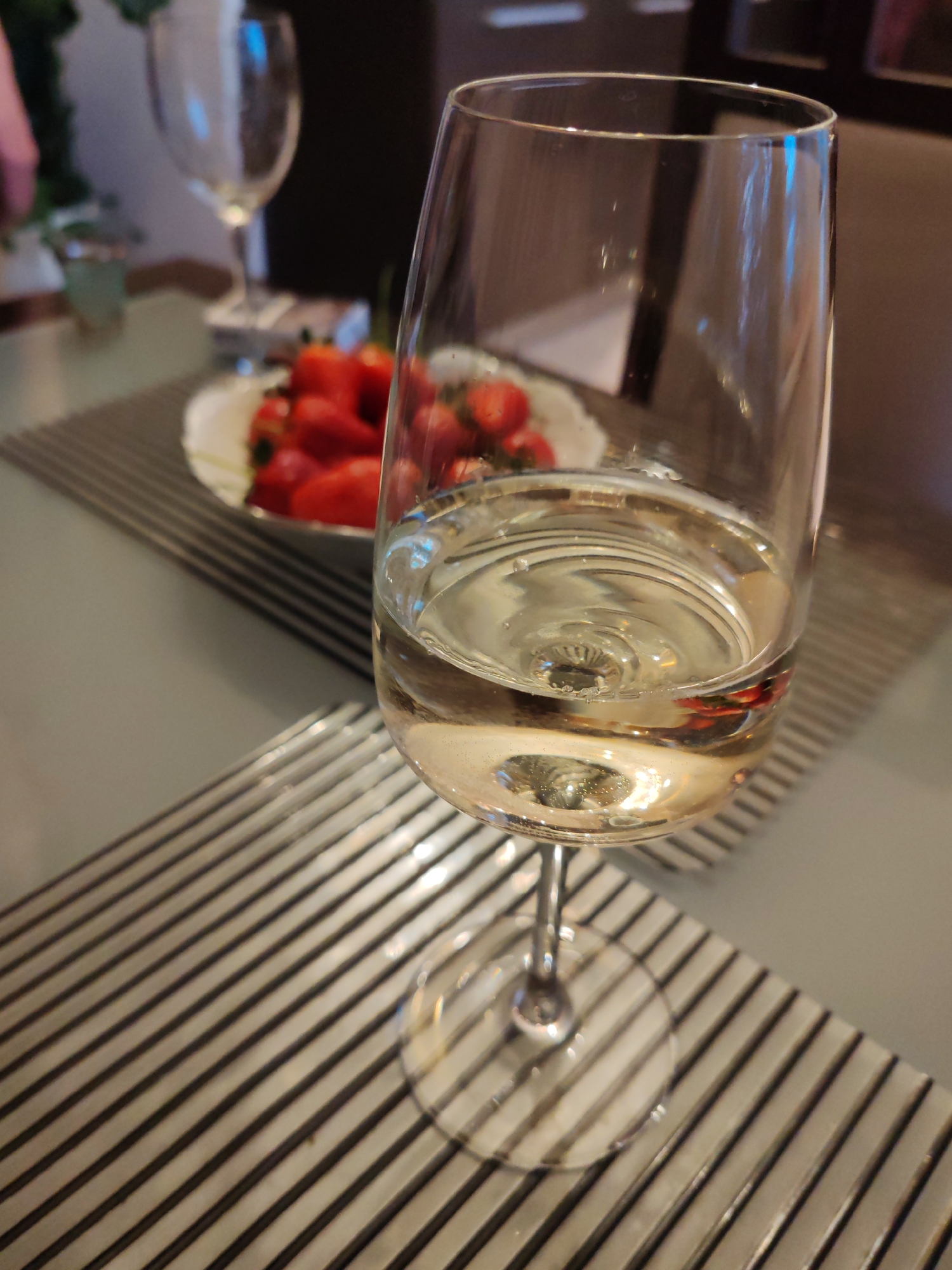 A close-up shot depicts a wine glass filled with a light-colored liquid, possibly white wine, standing on a gray-toned surface with dark stripes. Behind the wine glass, a plate containing strawberries is visible, slightly out of focus. In the background, another wine glass is present, along with indeterminate objects and indistinct outlines. The setting appears to be indoors, perhaps on a table. The image does not provide sufficient information to confirm the location as Saint Petersburg, Russia.