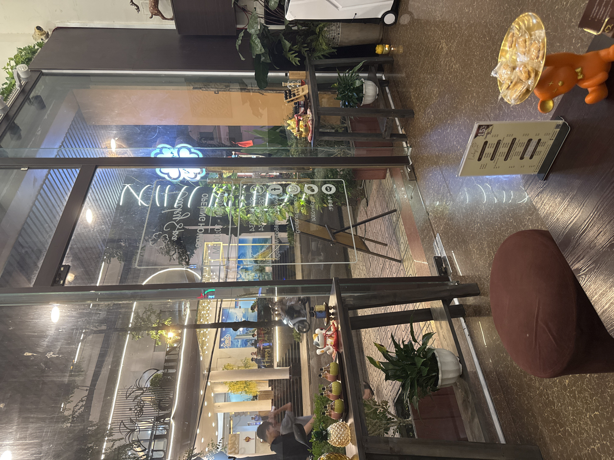 The image captures the exterior view of a business in Nha Trang, Vietnam, likely a spa or wellness center, as indicated by the neon sign that reads "OPENING HOURS" and "MASSAGE". The establishment features large glass windows that allow a view into the interior.
In the foreground, a wooden table displays decorative items including a pineapple-shaped glass object, a tray with several small bottles, and a "maneki-neko" or "beckoning cat" figurine. To the right, another wooden table holds a golden abacus, small bottles, and a potted plant.
Through the glass, the interior of the establishment is visible, with plants and lighting contributing to a serene ambiance. A white scooter is parked outside the entrance. Further back, a building with a modern facade and illuminated signage can be seen. A group of people are seated at tables on an elevated platform, suggesting an outdoor seating area or entrance.
The street outside appears to be well-lit, with reflections visible on the wet pavement, suggesting recent rain. The overall scene depicts a tranquil and inviting atmosphere.