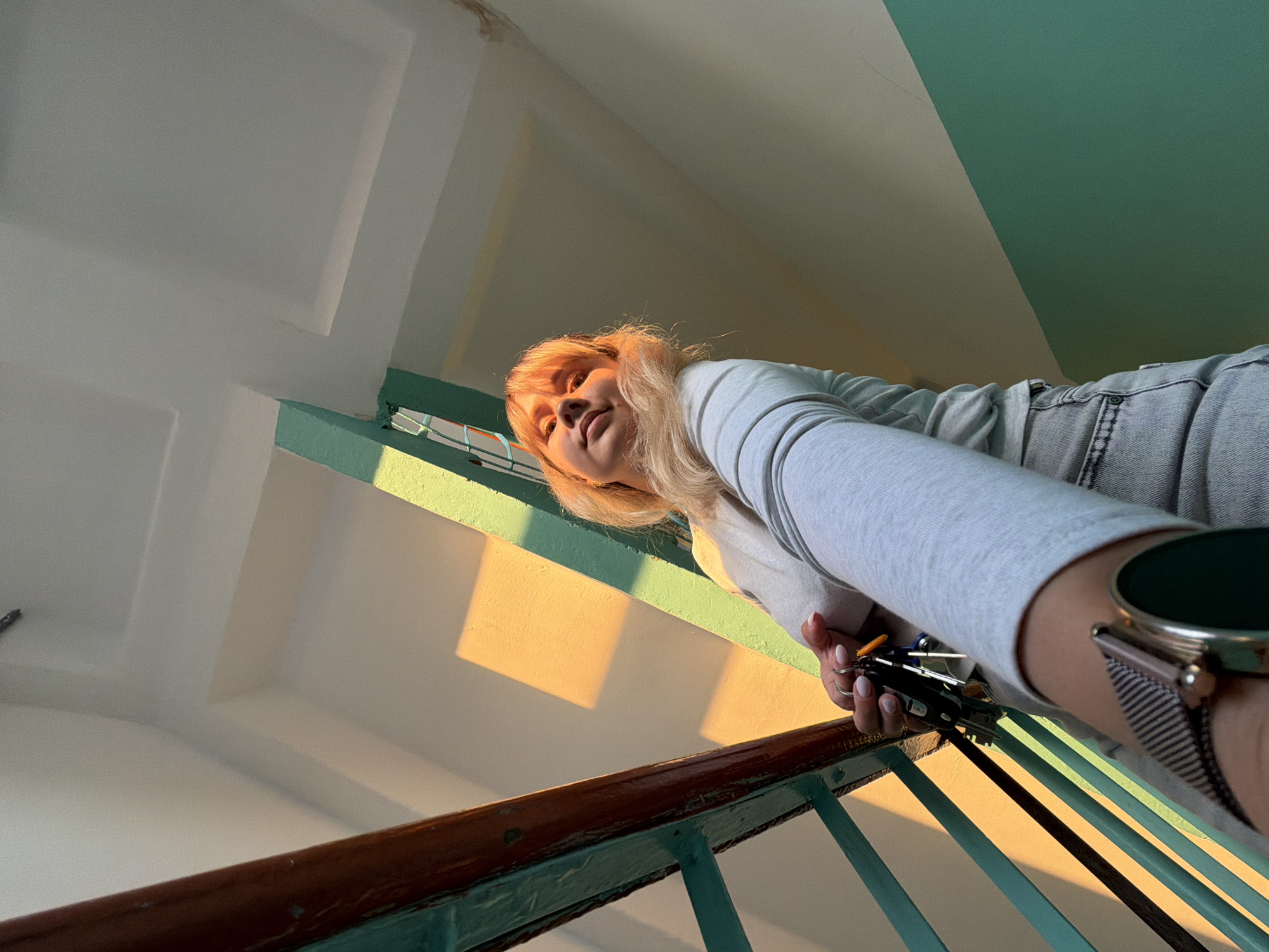 A young woman with blonde hair and bangs is captured from a low-angle perspective on a staircase, likely within a building in Chaikovsky, Russia. She is looking directly towards the camera, leaning slightly over a staircase railing. She wears a light gray long-sleeved top and light blue jeans. On her left wrist, she has a smartwatch with a metal mesh band, and in her right hand, she holds a set of keys.
The setting is an indoor staircase characterized by light-colored walls, possibly white or light beige, contrasted with sections painted in a distinct teal-green color. The railing has a brown wooden handrail atop teal-green vertical bars. Strong, warm, golden sunlight illuminates the scene from a low angle, casting prominent, elongated shadows on the walls and floor, suggesting either early morning or late afternoon (golden hour). The directional light highlights her face and hair. Her expression is calm and direct. There is no clearly visible text in the image.