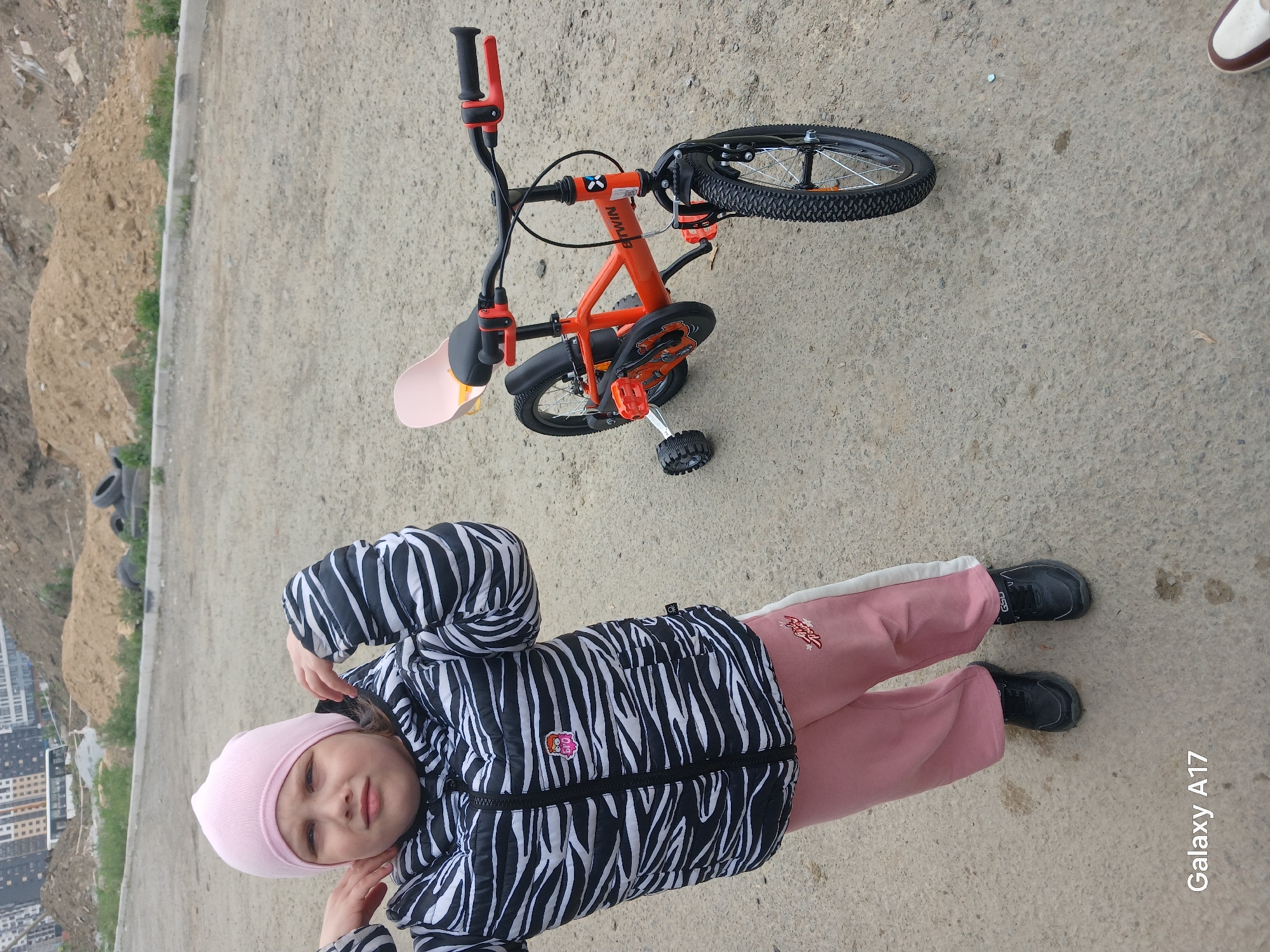 A young child in a pink hat, zebra-striped jacket, and pink pants lies on a concrete surface, seemingly a sidewalk or road. Next to her is a small, orange child's bicycle with training wheels. The setting appears to be a construction site or undeveloped area on the edge of a city, with a pile of dirt and some old tires visible in the background. There are also some buildings visible in the distance, potentially located in the city of Yekaterinburg, Russia. The photo was taken with a Galaxy A17 phone.