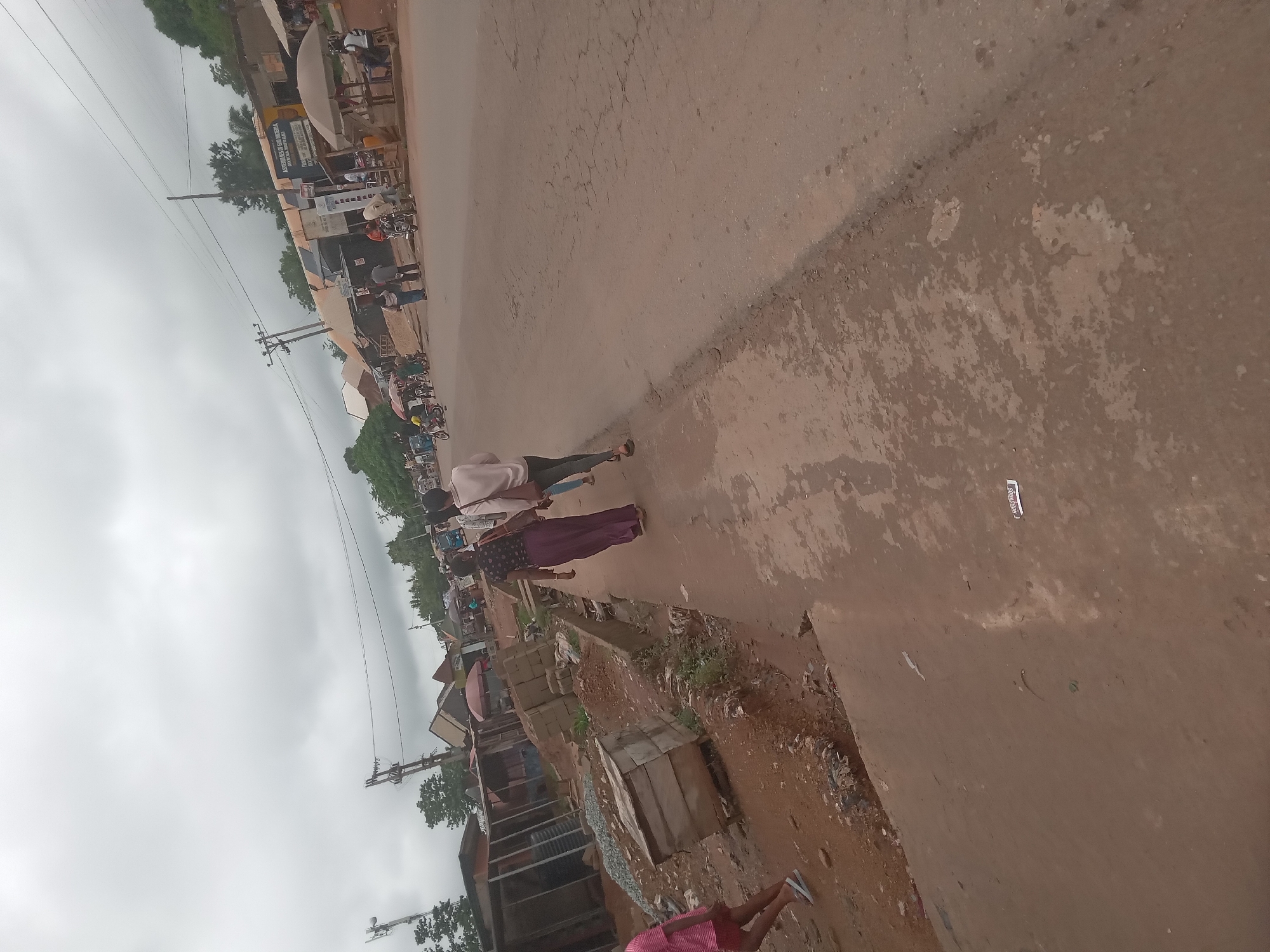 The image shows a street scene in Achara Ndiagu Omegio, Nigeria. The street is unpaved and appears muddy or dusty. Several people are walking along the road. Buildings and shops line one side of the street. Overhead power lines are visible. There are also vehicles and motorbikes parked on the side of the road. The sky is overcast. The image appears to have been taken at ground level.