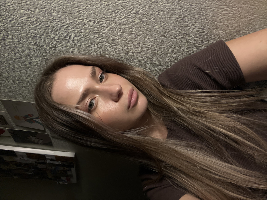An indoor portrait depicts a single female subject positioned against two distinct wall surfaces. The individual, identified as a young adult female, has long, straight to slightly wavy light brown hair with blonde highlights, light-colored eyes, and pink lips. She wears a dark brown, ribbed, short-sleeved top. Her expression is neutral, and her gaze is directed generally towards the camera.
The background on the subject's right (viewer's left) consists of a light grey or beige wall with a distinct, vertical, wave-like textured pattern, possibly wallpaper. To the subject's left (viewer's right), the background transitions to a darker, untextured wall surface or a shadowed corner. Affixed to this darker area are several items: a white-framed board displaying multiple small photographs or images, and additional unframed prints or postcards, including one clearly illustrating "The Little Prince."
Lighting in the scene is uneven, suggesting a primary light source positioned to the subject's right (viewer's left), illuminating her face and the textured wall prominently. The overall environment is consistent with a private residential interior. No specific architectural features, landmarks, or other geographical markers are present to identify a precise location within the city of Saint Petersburg, Russia.