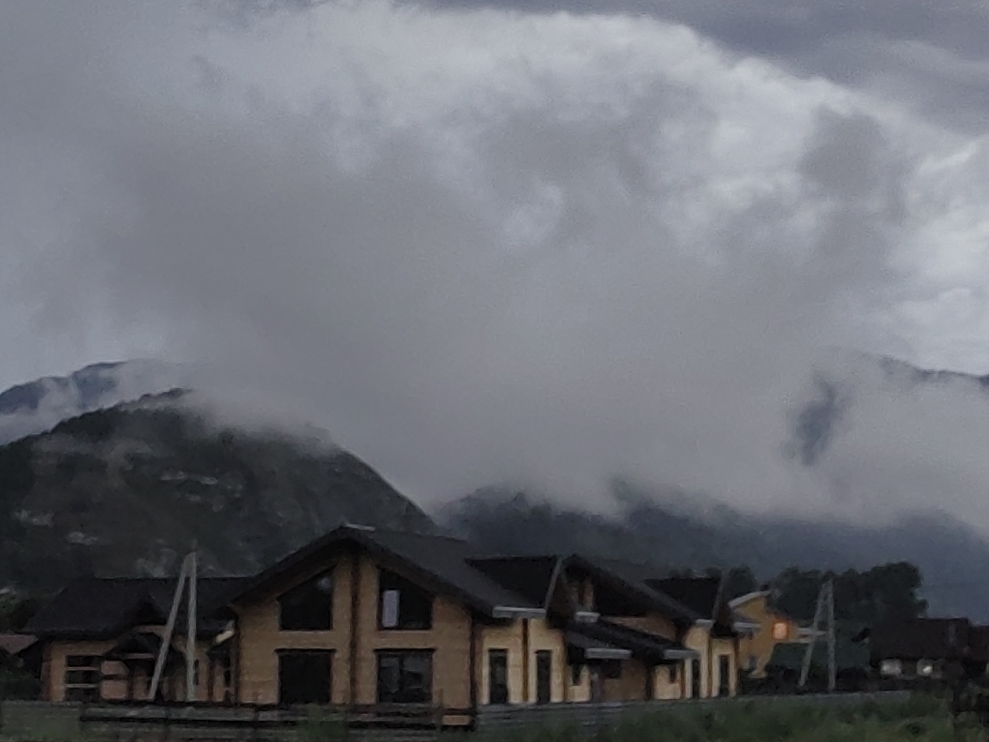 The image depicts a row of modern wooden houses situated in a rural mountainous landscape, likely in Anos, Russia. The houses feature dark roofs and light-colored wooden exteriors with large windows. The sky is overcast and filled with dense, gray clouds, with a portion of a dark, mist-shrouded mountain visible in the background. The overall atmosphere is somber and cloudy, suggesting a cool or possibly rainy day. There are no people visible in the image.