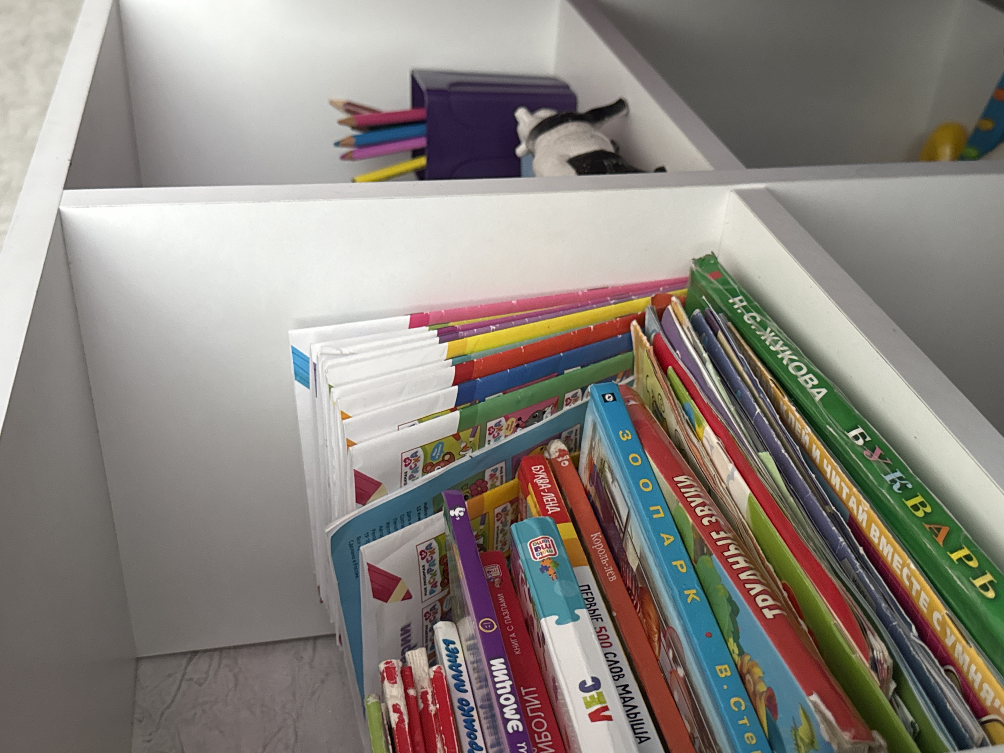 A close-up shot shows a white shelf unit filled with books and art supplies. In the foreground, a stack of colorful children's books leans to the left. The titles are in Russian and include "ГРОМКО ПЛАЧЕТ," "ЭМОЦИИ," and "ИБОЛИТ." To the right, on the same shelf, a purple cup holds several colored pencils, and a small black and white cow figurine stands on its hind legs. The shelf unit is positioned in an indoor environment.