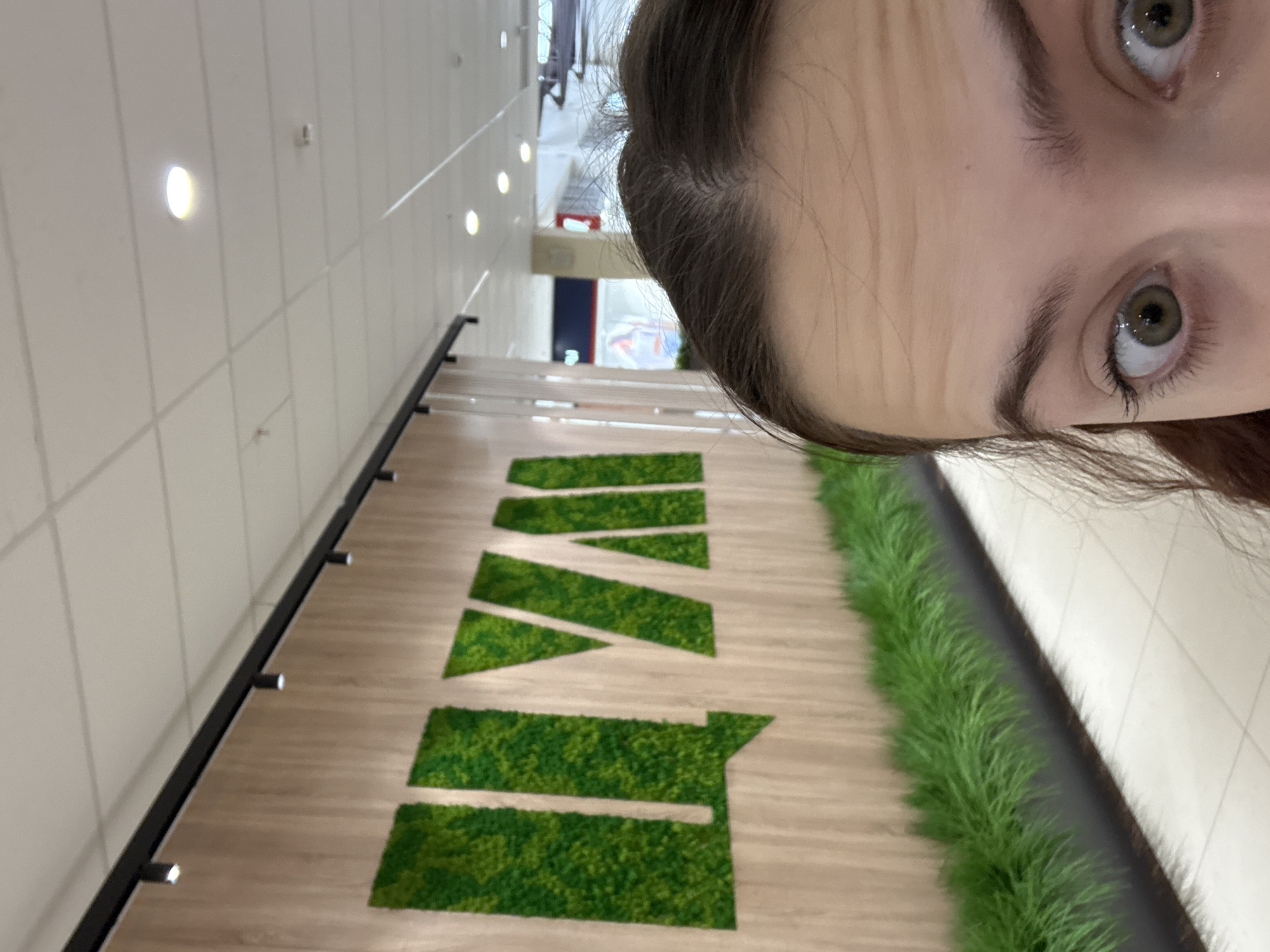 The image shows a person in an indoor environment, likely a commercial or public space in Unknown, Unknown. The primary focus is on the person's face, specifically the eyes and forehead, positioned at an angle relative to the background. The background features a wall with evenly spaced, recessed lights, and a floor design with large green letters spelling out what appears to be "IVE." The letters are composed of a green moss-like material. Additionally, there's a strip of longer green grass-like material running alongside the letters. The overall setting seems to be modern, possibly a retail store, exhibition space, or office area.