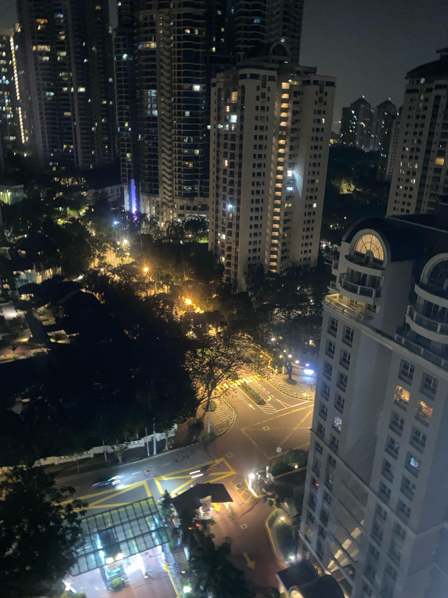 This image captures a nighttime aerial view of a city intersection in Kuala Lumpur, Malaysia, likely in a densely populated urban area. Tall, modern skyscrapers dominate the background, their windows illuminated with a warm, golden light. In the foreground, a multi-lane road curves through the scene, marked with yellow lines and pedestrian crossings. Several cars are visible on the road, their headlights and taillights streaking due to the long exposure, indicating movement. Trees with dark foliage line the sides of the road, some with streetlights casting a yellow glow. A large, modern building with a distinctive arched window detail stands prominently on the right side of the frame. The overall impression is one of a vibrant, bustling city at night.