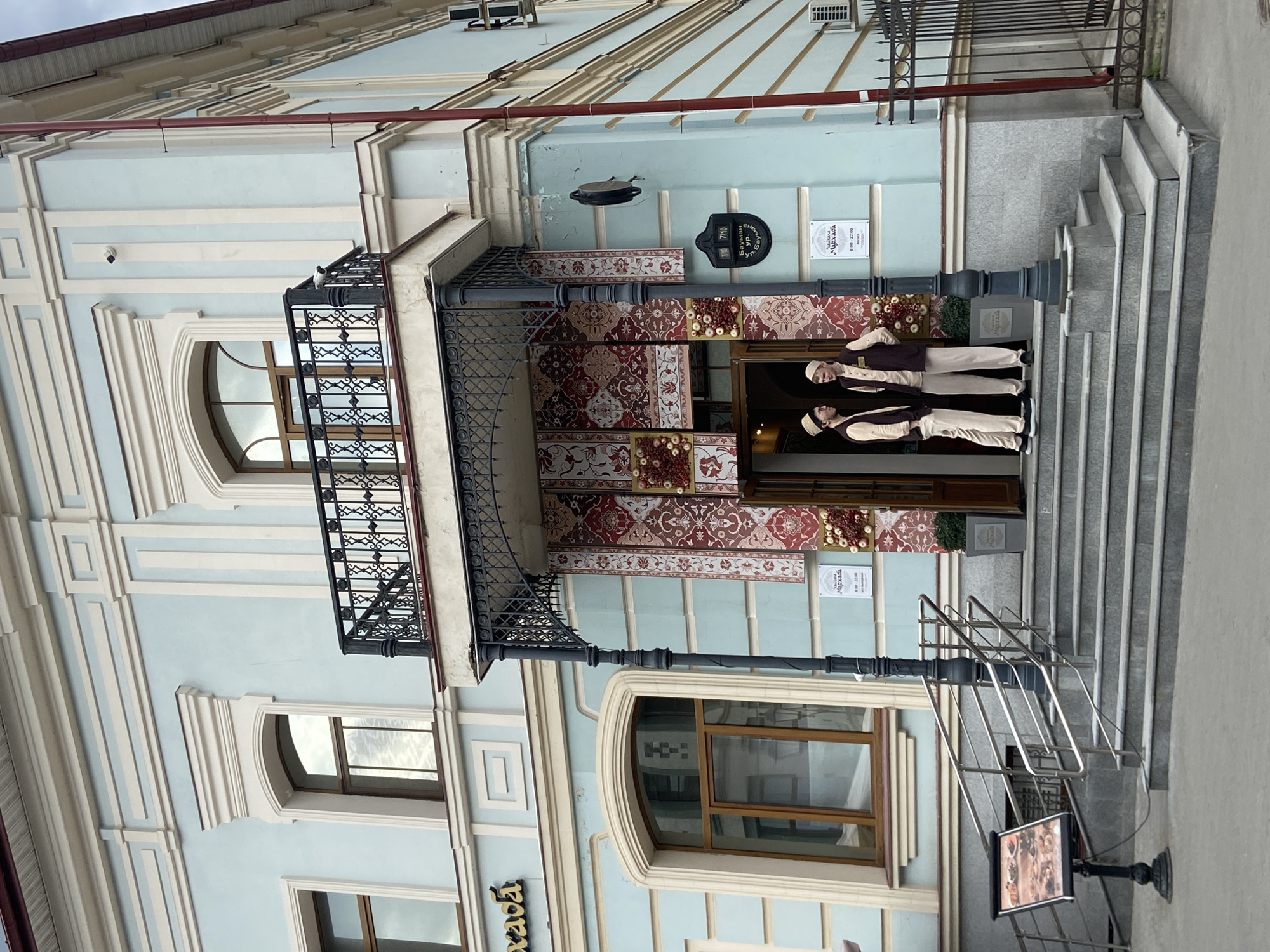 The image depicts the facade of a building in Kazan, Russia. The building is light blue with white trim and several windows. A balcony with wrought iron railings is visible on the second floor. The entrance is decorated with carpet-like patterns and flanked by two mannequins dressed in traditional Tatar clothing. Signage in both Russian and another language (possibly Tatar) adorns the building's facade. A sign with the name "Мархаба" and the hours of operation "8:00-22:00" is also present. There is a small menu board on the right. The building is accessed by a set of stairs.