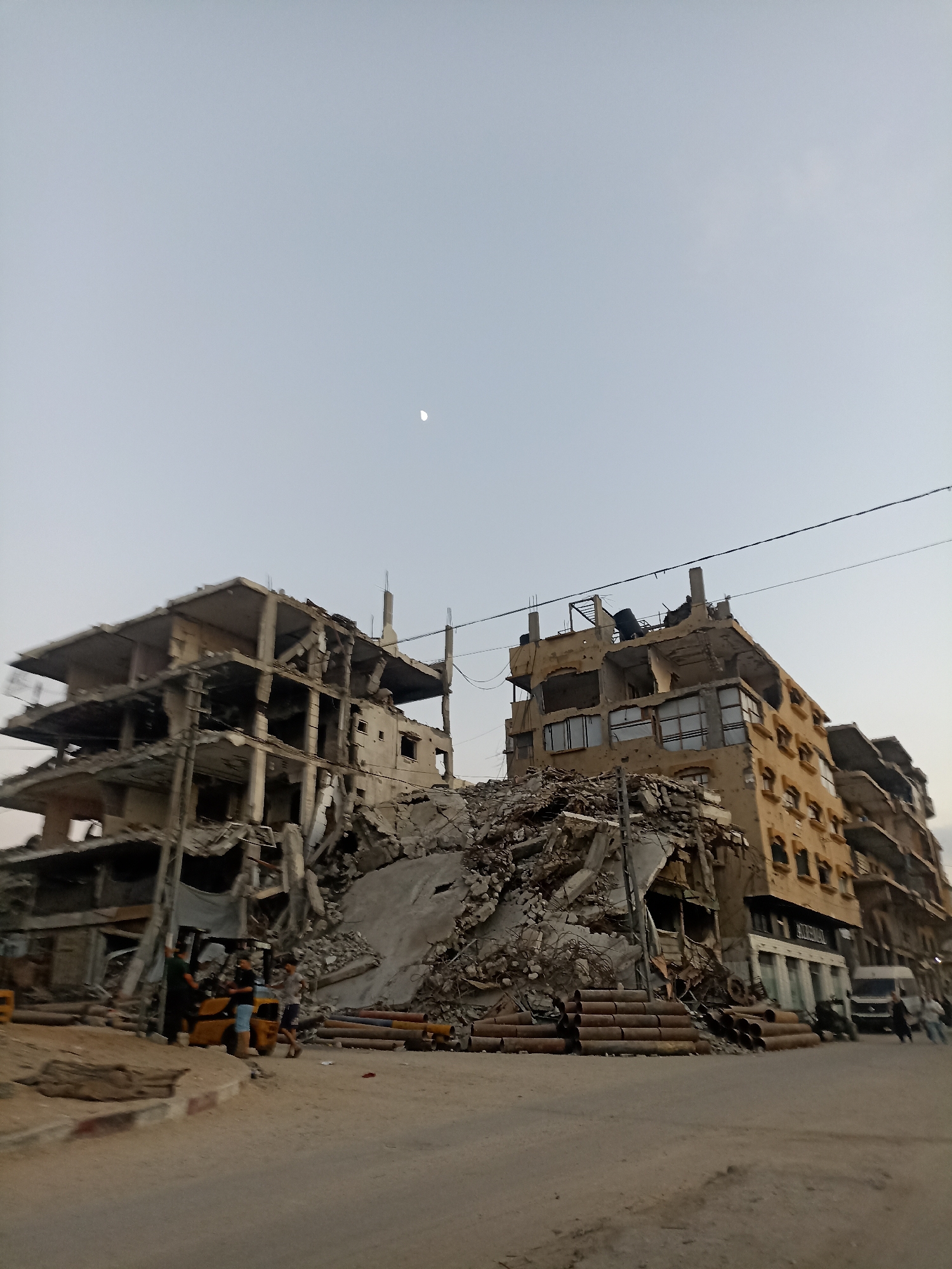 The image depicts a street scene in Gaza City, Palestinian Territories, characterized by extensive destruction of buildings. Two multi-story concrete structures are heavily damaged, with exposed rebar and collapsed sections. One building on the left appears to be a partially collapsed concrete frame. To the right, a multi-story building with a yellow facade shows significant damage to its upper floors, with broken windows and missing sections. A large pile of rubble lies in the foreground, obscuring the base of the buildings.
In the mid-ground, a yellow forklift is parked on the dirt road, with three individuals nearby. One person is standing by the forklift, while two others are interacting. A white van and several people are visible further down the street to the right, suggesting ongoing activity or movement. Large cylindrical objects, possibly pipes, are stacked on the ground near the rubble. Overhead, power lines stretch across the frame, and a sliver of the crescent moon is visible in the pale sky. The overall impression is one of devastation and the aftermath of conflict.