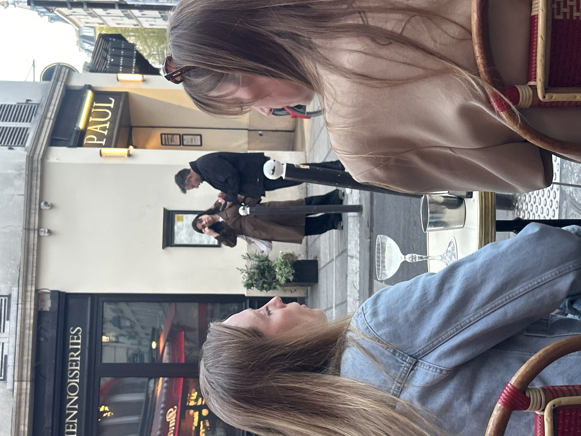 Two women are seated at an outdoor cafe. One is wearing a beige jacket and sunglasses, the other a denim jacket. On the street, a man in a black coat and a black cap is talking to a woman in a brown coat who is holding a phone. Behind them is a "PAUL" bakery. Across the street are the words "IENNOISERIES". There are glasses on the cafe table. The cafe chairs are red and wicker.
