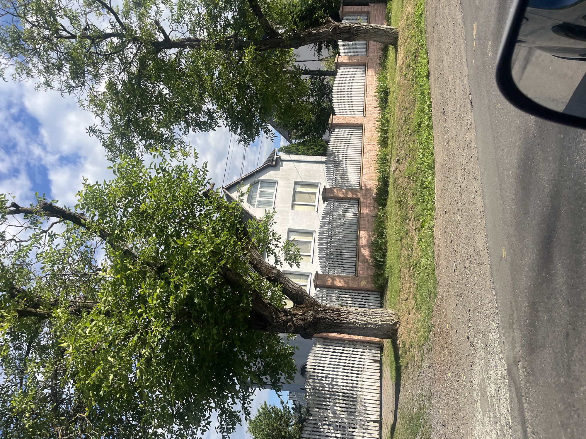 A medium-shot image captured from a low angle shows a residential house with a white picket fence in front of it. The house is a two-story building with a gray facade and white-framed windows. The fence is constructed of white wooden pickets and is topped with a light pink brick base. Lush green trees with dense foliage are positioned in front of the fence, partially obscuring the house. The ground in front of the fence consists of a gravel path and a strip of green grass. A paved road is visible in the foreground, with a car's side mirror reflected in the lower right corner of the image. The sky is blue with scattered white clouds. The scene depicts a typical suburban or rural setting in the city of Storozhnytsia, Ukraine.