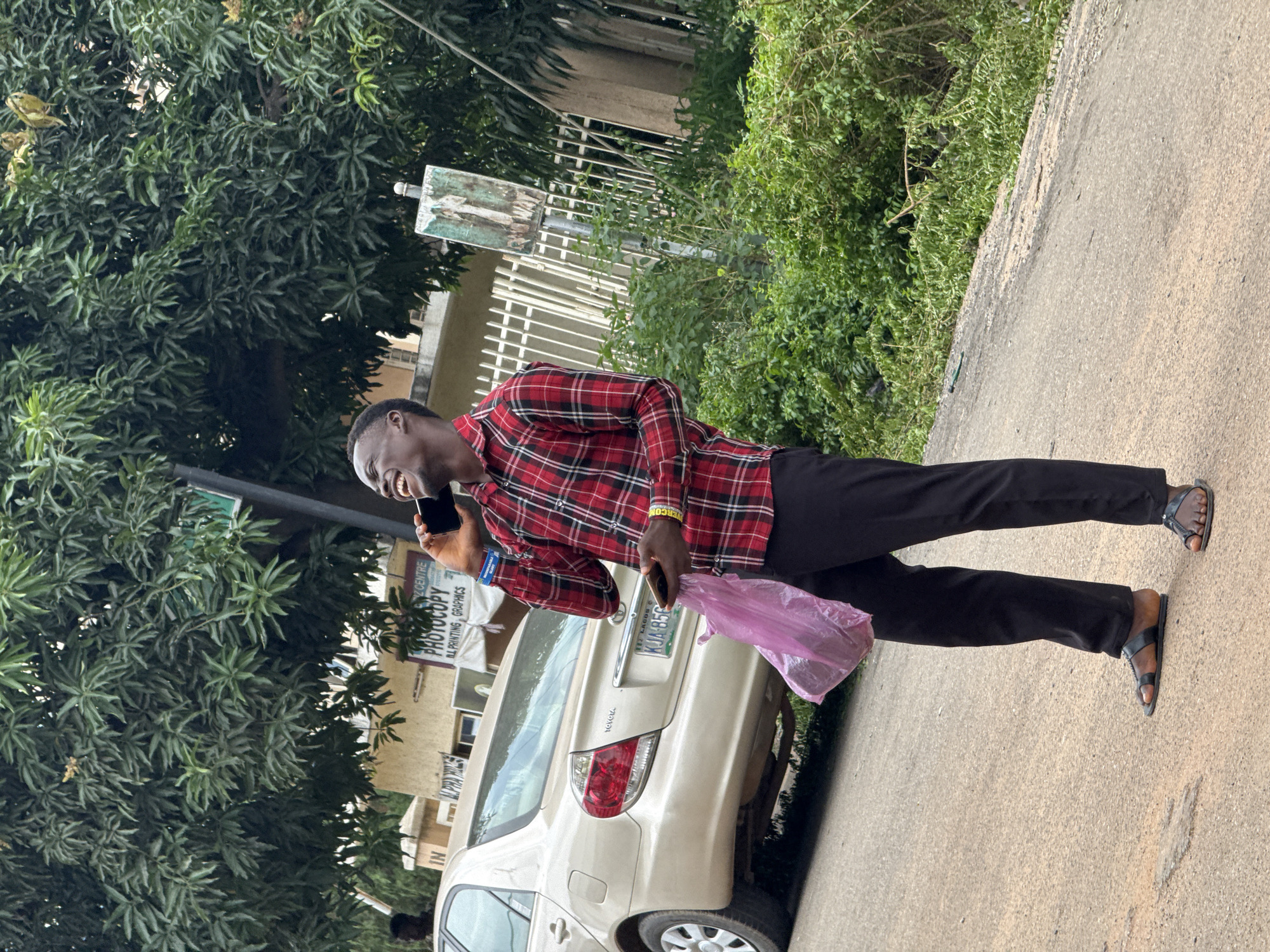 A young Black man is standing on a paved road next to a beige Toyota Camry. He is wearing a red and black checkered long-sleeved shirt, black pants, and black sandals. He is laughing and holding a dark-colored smartphone to his right ear, and a pink plastic bag in his left hand. The car has a license plate that reads "KJA 856". In the background, there is lush green foliage, a building with a "PHOTOCOPY" sign, and a white metal fence. The scene appears to be in an urban area of Abuja, Nigeria.