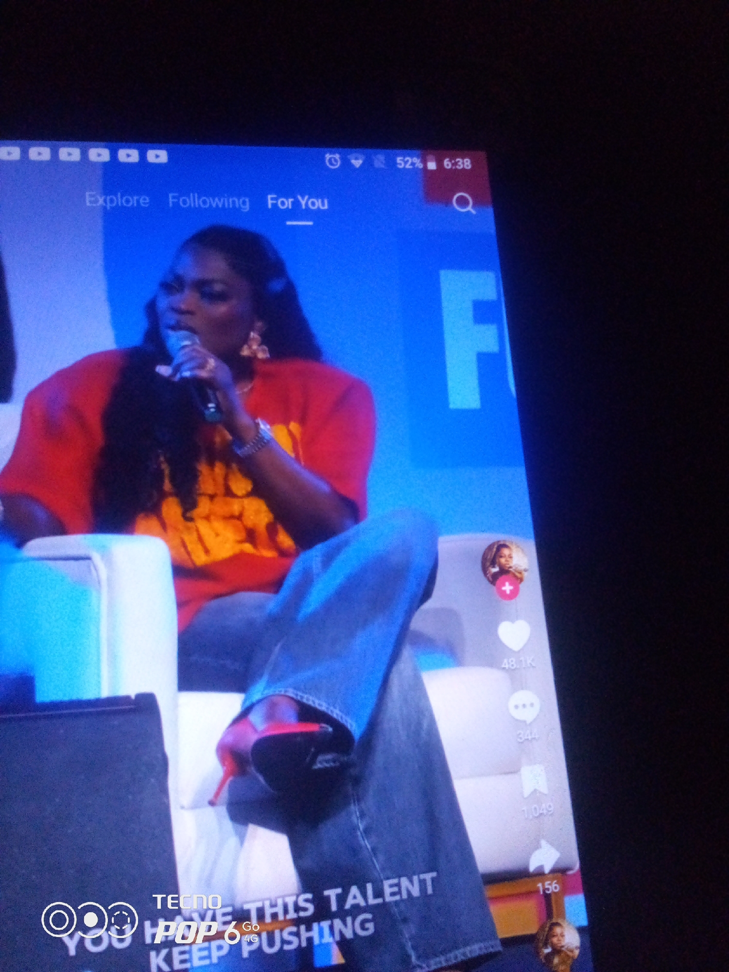 The image displays a screen, likely a smartphone or tablet, showing a video of a woman with dark, curly hair singing into a microphone. She is wearing a bright red t-shirt with yellow lettering that is partially obscured. Her attire also includes blue jeans and red high-heeled shoes. She is seated, and part of a white chair is visible to her left.
Overlaying the video are various user interface elements, typical of a social media platform. Along the top, there are icons for "Explore," "Following," and "For You." To the right, the screen indicates a battery level of 52% and the time as 6:38. There's also a search icon. On the right side of the screen, there are engagement metrics: a profile picture with a red plus icon, a heart icon with "48.1K" next to it, three dots icon with "344," a bookmark icon with "1,049," and a share icon with "156."
In the lower left corner, a watermark reads "TECNO POP 6 GO" and another line of text, partially visible, includes "YOU HAVE THIS TALENT KEEP PUSHING."
The context of this image does not provide any information about the city of Howayi, Nigeria. The scene is entirely contained within the digital display of a device.