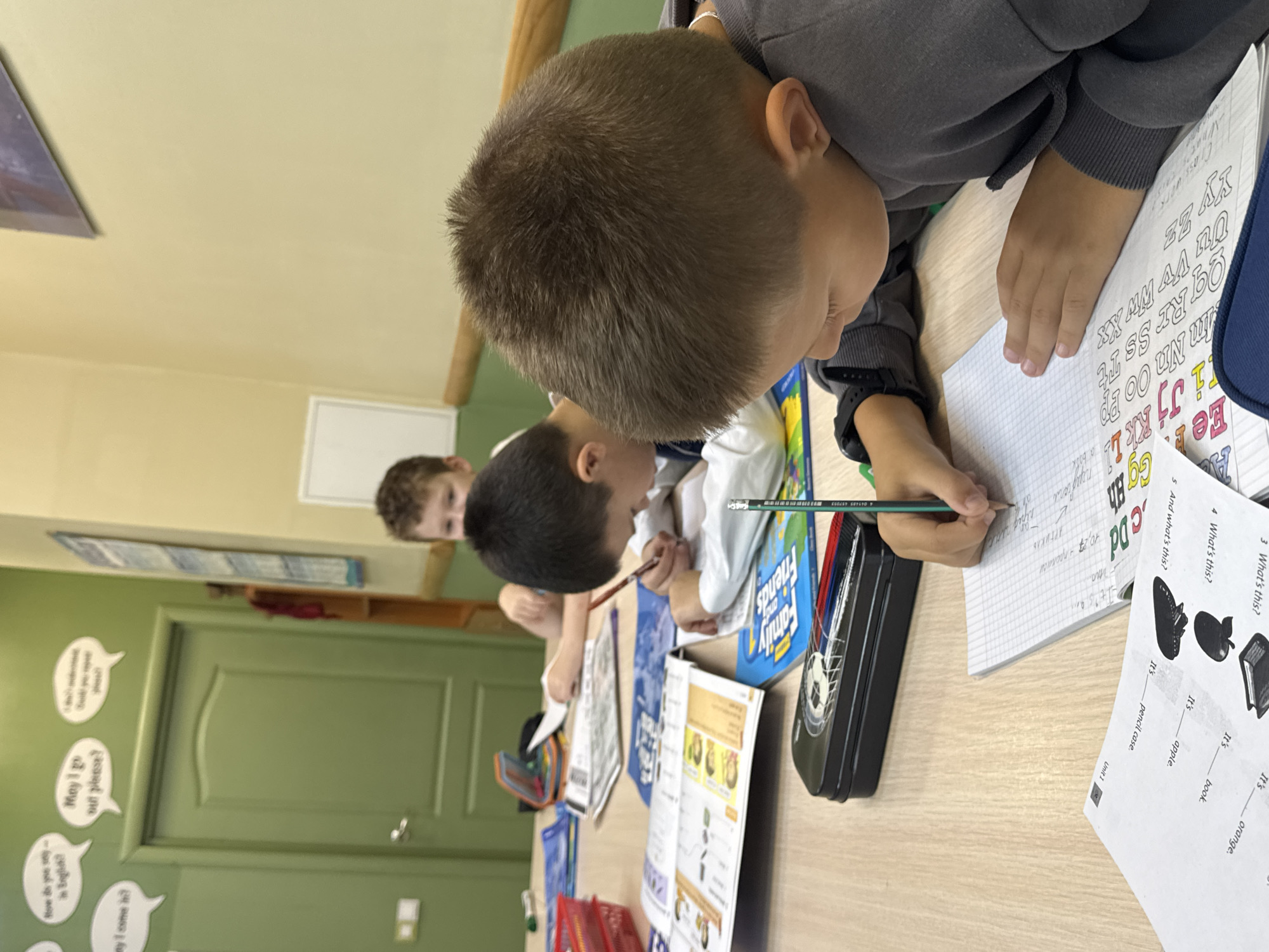 The image shows three young boys sitting at a wooden table, engaged in schoolwork. The boy in the foreground is right-handed, writing in a notebook with a pencil. He wears a gray hooded sweatshirt and a black watch. A black pencil case with a soccer ball design is in front of him. To his left, another boy, also right-handed, is writing in a notebook with a pencil. He wears a white shirt. Further back, a third boy, wearing a light blue shirt, is also focused on his work, with his arms resting on the table.
The table is covered with books, notebooks, and writing utensils. On the wall behind the boys, there are speech bubbles with English phrases like "May I go out please?" and "How do you say... in English?". There is also a door painted green and a whiteboard. The overall environment appears to be a classroom or a study area.
Based on the presence of English language learning materials and the style of the room, this scene likely depicts an English language lesson at a private educational institution or a specialized language school. The image does not contain enough information to pinpoint a specific location in Perm, Russia.