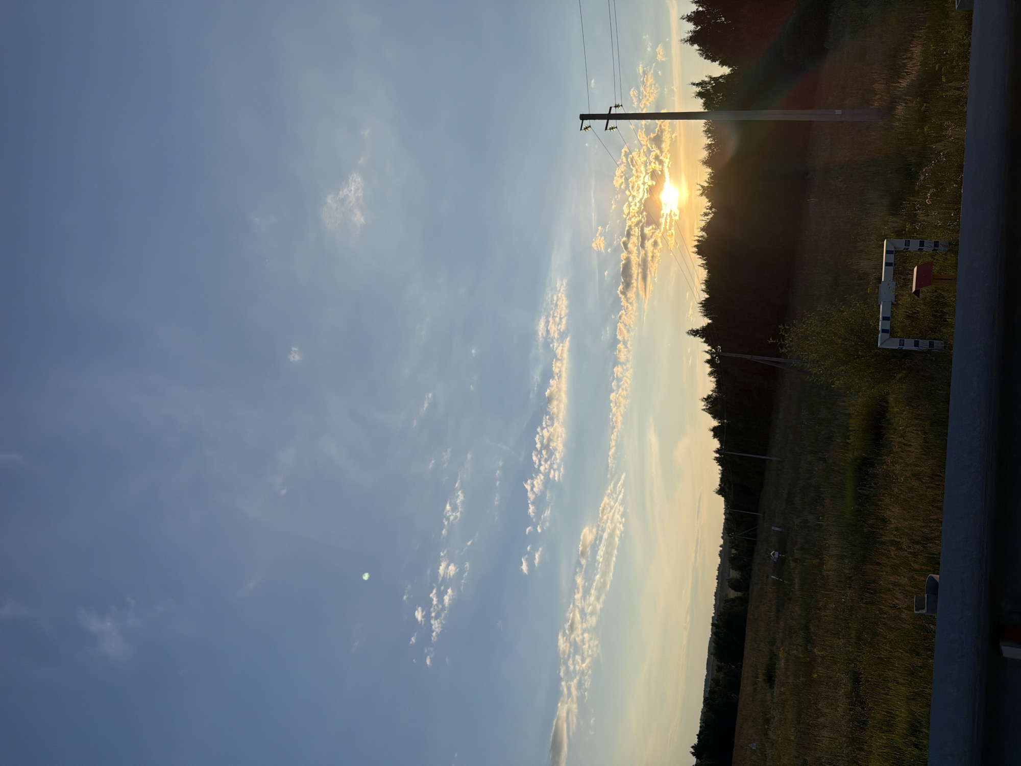 The image captures a rural landscape in Obukhovo, Russia, during sunset. The sky is a vibrant blue with scattered, wispy clouds tinged with orange and yellow hues from the setting sun. The sun itself is low on the horizon, partially obscured by a band of clouds, and its rays are casting a warm glow across the scene.
In the foreground, a grassy field stretches out, appearing somewhat wild with tall grasses and scattered wildflowers. A dark line of trees forms a dense forest along the distant horizon. A solitary utility pole with power lines stands prominently on the right side of the frame, silhouetted against the bright sky. The pole has insulators and wires extending from it. A low, grey barrier runs along the very bottom of the image, possibly part of a road or viewing platform. In the middle ground, there are a few small, white objects in the field, possibly markers or signs, and a small, red mailbox-like structure with a white and blue striped frame. The overall impression is one of natural beauty and a serene, rural atmosphere.