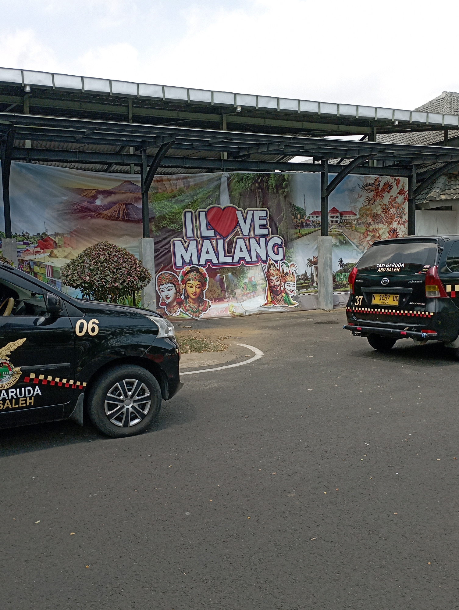 The image depicts a street scene in the city of Pakis, Indonesia. In the foreground, two black cars are parked on an asphalt road. The car on the left, a minivan, has the number "06" in gold lettering on its side and features branding that includes "GARUDA SALEH". The car on the right, also a minivan, has the number "37" and a license plate reading "N 1457 UG". A banner displaying "I LOVE MALANG" is prominently featured in the background, along with images of volcanoes, traditional dancers, and batik patterns. The banner is suspended under a covered walkway structure with metal beams and translucent roofing. A dense, bushy tree is situated in front of the banner, between the two cars. The sky is overcast.
