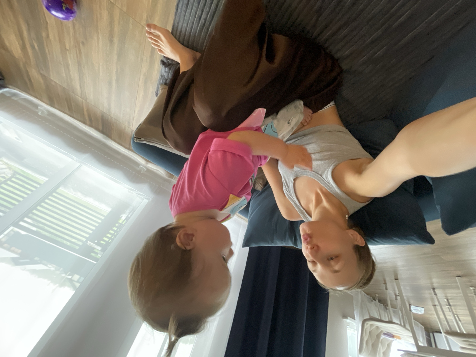 A woman with blonde hair pulled into a bun and a gray tank top is sitting on a dark blue couch. She is puckering her lips towards a baby who is sitting on her lap. The baby is wearing a pink shirt with colorful designs. The woman's right arm is extended towards the camera, holding a phone. The baby's left foot is visible, peeking out from under the woman's brown pants. A light blue and white object, possibly a wipe or diaper, is held in the woman's hand, near the baby. The scene appears to be indoors, with a wooden floor and a large window with white curtains in the background. The location is not specified as being in the city of Подшивалово, Russia.
