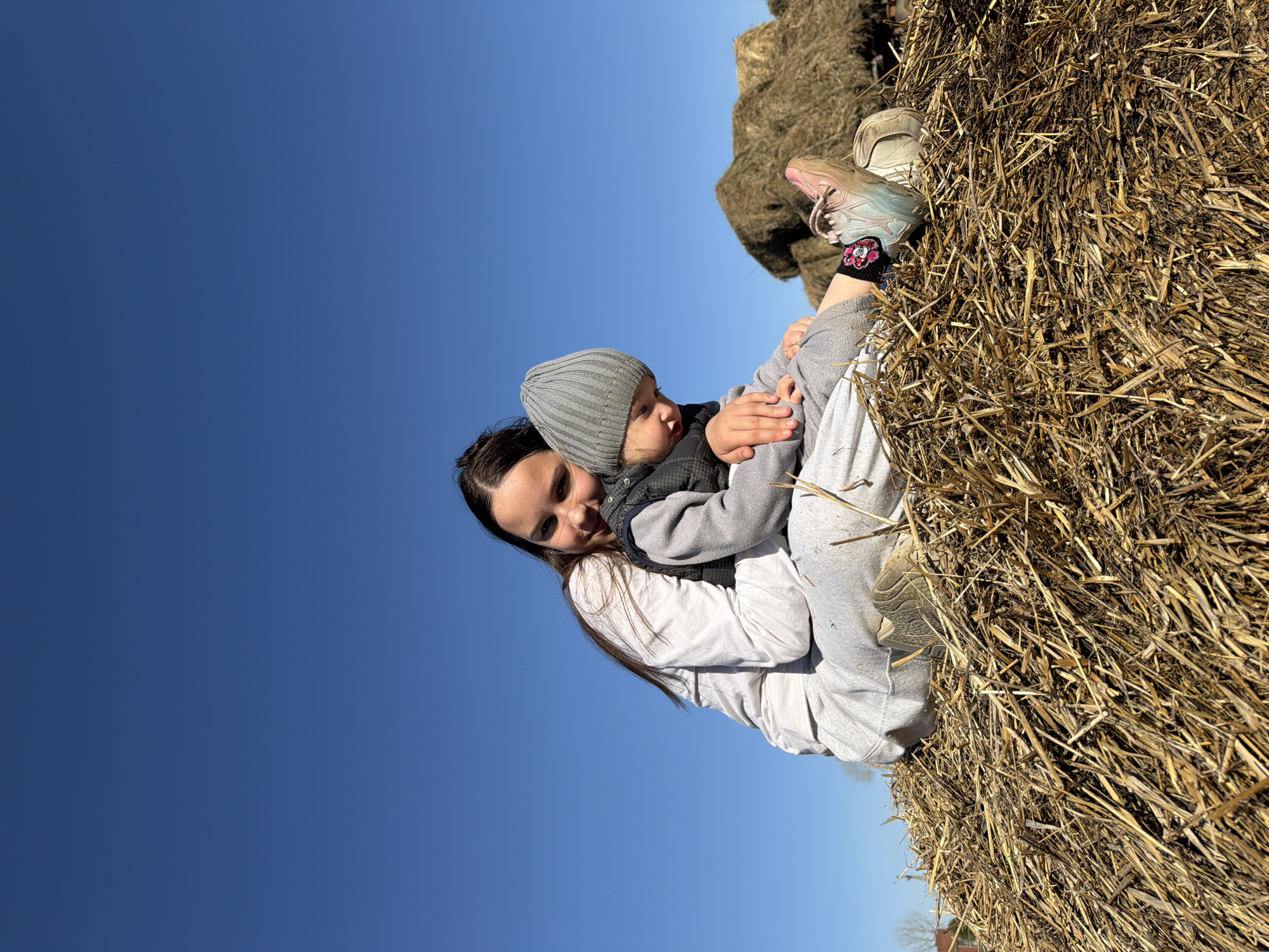 A woman and a young child are sitting on a hay bale. The woman has long, dark hair and is wearing a white shirt and gray sweatpants. The child is wearing a gray hat, a gray vest, and gray sweatpants. The child is sitting in the woman's lap and she is holding him. They are outdoors on a sunny day. The sky is blue and cloudless. The photograph was taken in Chaikovsky, Russia.