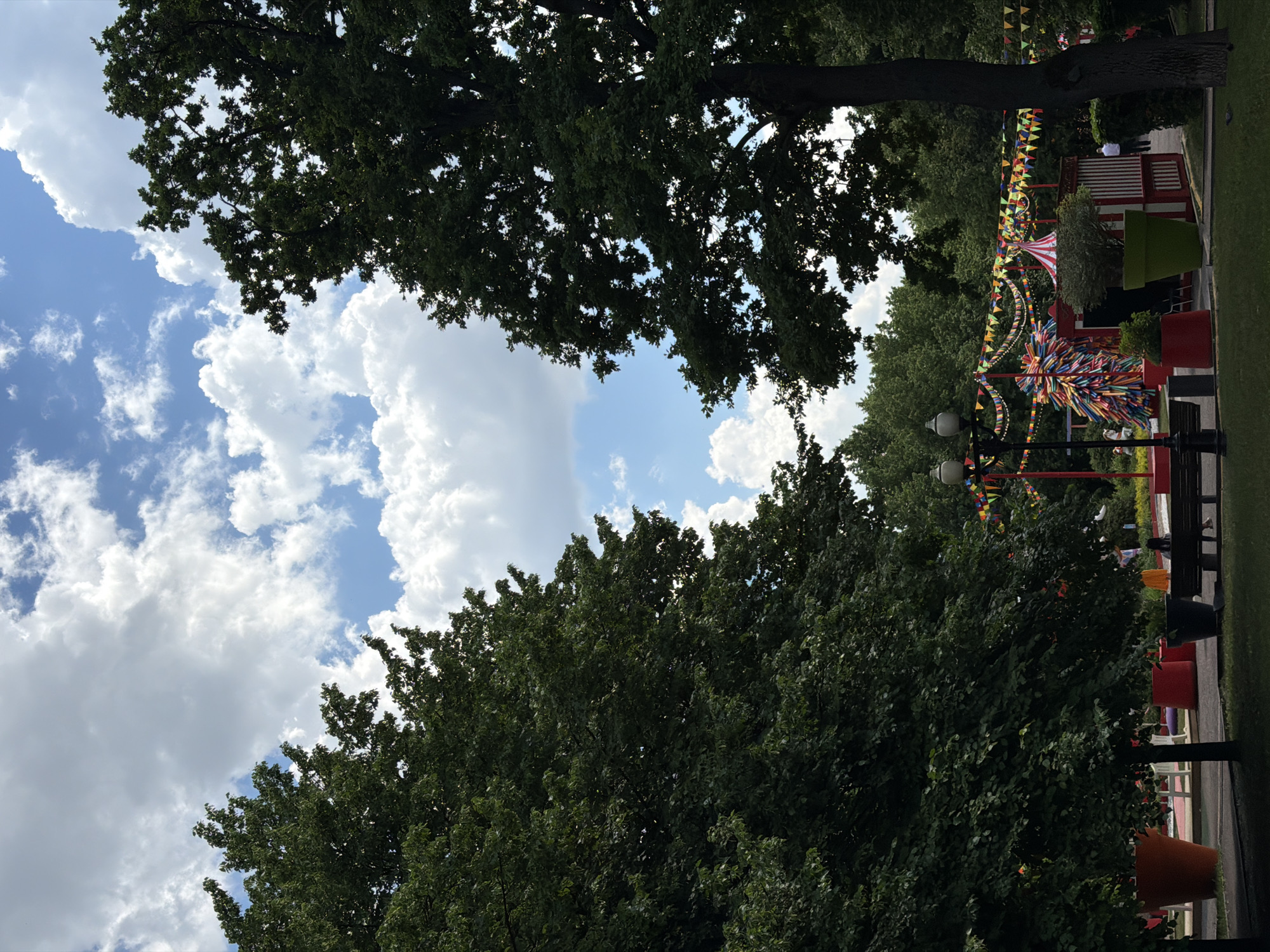The image captures an outdoor scene, likely a park or public recreational area, adorned with festive decorations under a bright, cloudy sky. The dominant features are several large trees with dense green foliage, framing the sky and partially obscuring the ground level.
The sky is a clear blue, filled with scattered, voluminous white cumulus clouds, suggesting fair weather during the daytime, possibly late morning or early afternoon.
Below the treeline, a vibrant, colorful setting unfolds. Numerous strings of triangular flags (bunting) in various hues crisscross the area, stretching between poles and structures. A large, multi-colored, pom-pom-like decoration or sculpture, resembling a burst of ribbons, is prominently featured. Nearby, a red and white striped conical structure, possibly a tent or part of a booth, adds to the carnival-like atmosphere. The ground area also features large, brightly colored planters in red, orange, green, and black, as well as dark-colored benches. Two street lamps or light fixtures are visible.
While no specific activity is clearly depicted, the abundant decorations strongly suggest an ongoing or recently set up festival, fair, or celebratory event. A few indistinct figures can be vaguely seen in the background, but no clear human activity or emotion is discernible. There is no visible text in the image. The overall mood conveyed by the bright colors and decorations is cheerful and lively.