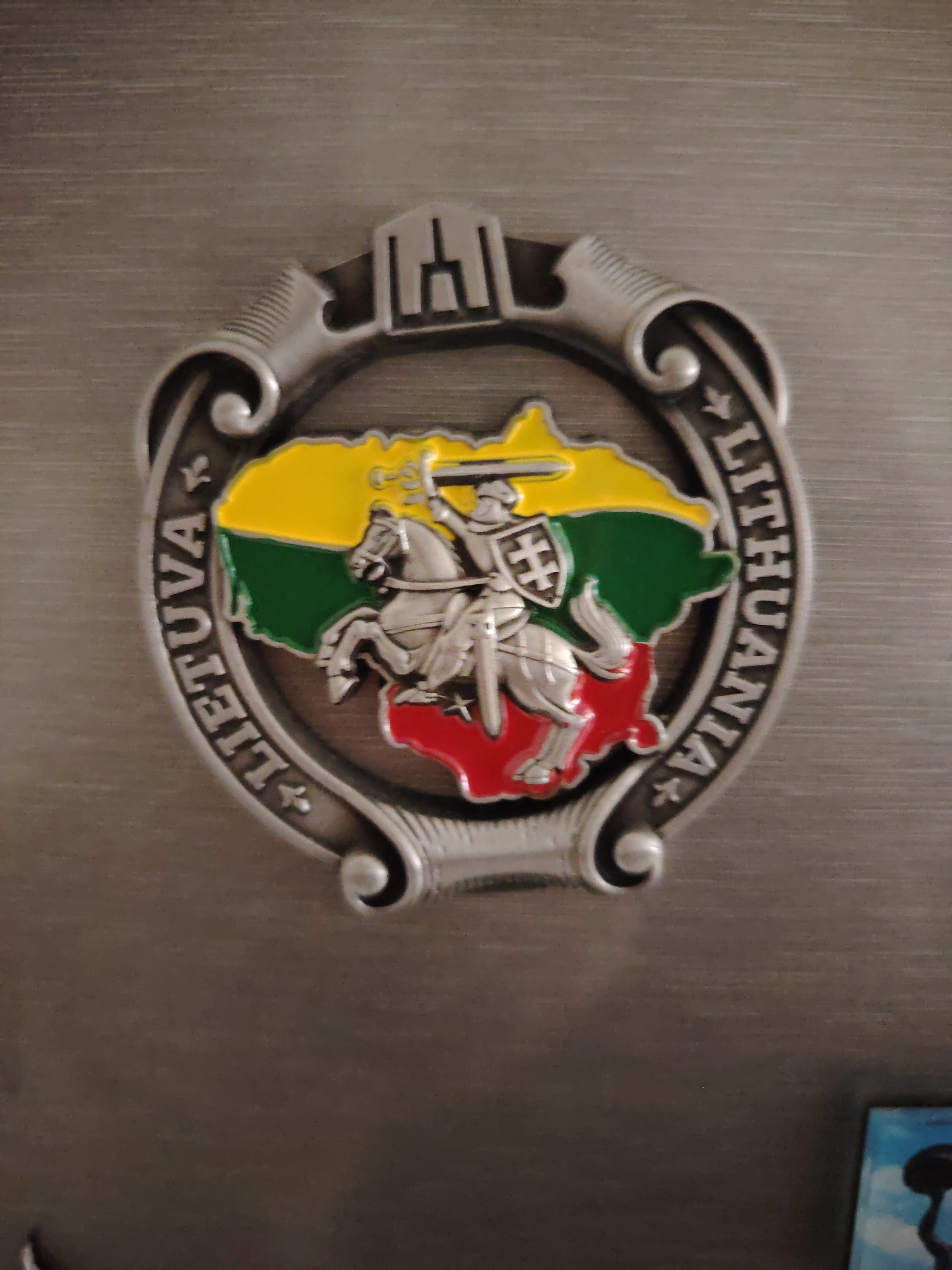 A metal relief magnet, depicting national symbols of Lithuania, is adhered to a brushed metallic surface within the city of Huntingdonshire, United Kingdom. The magnet is designed as an ornate, silver-colored frame with "LIETUVA" inscribed on the left arc and "LITHUANIA" on the right arc, separated by small stylized crosses. At the top of the frame, a stylized architectural structure, possibly a tower or castle, is featured. The interior of the frame contains a raised map outline of Lithuania, colored with enameled yellow (top), green (middle), and red (bottom) stripes, representing the Lithuanian flag. Superimposed on this map is a silver-colored relief of the Vytis, the national coat of arms of Lithuania: a knight on horseback with a raised sword in his right hand and a shield bearing a double cross in his left. The background surface exhibits horizontal graining. A small, indistinct blue and white object is partially visible in the bottom right corner of the frame.