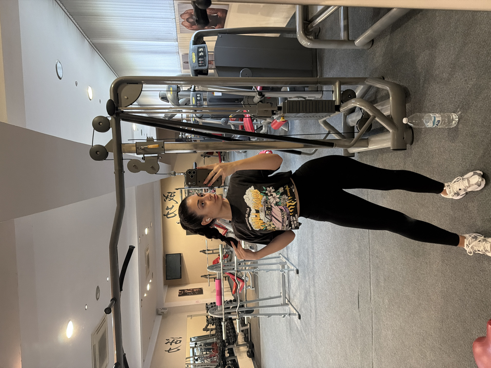 A woman stands in a gym, holding a phone and posing for a picture. She is wearing a black t-shirt and leggings. The gym features various exercise machines, weights, and mirrors. The location is likely in Blagoevgrad, Bulgaria.