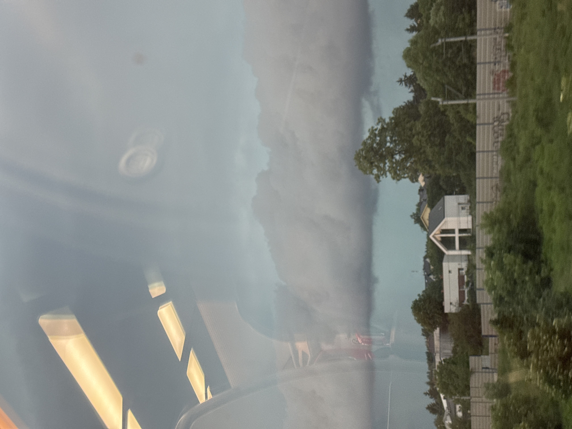 The image appears to be taken from inside a vehicle, possibly a train, looking out a window. Reflections on the window suggest interior lighting, with rectangular, illuminated panels visible in the upper left portion. The exterior scene depicts a suburban or semi-rural environment under a cloudy, possibly stormy, sky.
In the mid-ground, there are several houses, one of which is a modern, white structure with a distinctive A-frame roof. Trees are scattered throughout the landscape, with denser foliage forming a line along the mid-ground, behind a fence. The fence appears to be a noise barrier, common along transportation routes.
In the foreground, there is a grassy area with more greenery. The overall atmosphere is somewhat somber due to the heavy cloud cover. There are no visible people or specific actions taking place. The location is identified as Luboń, Poland.