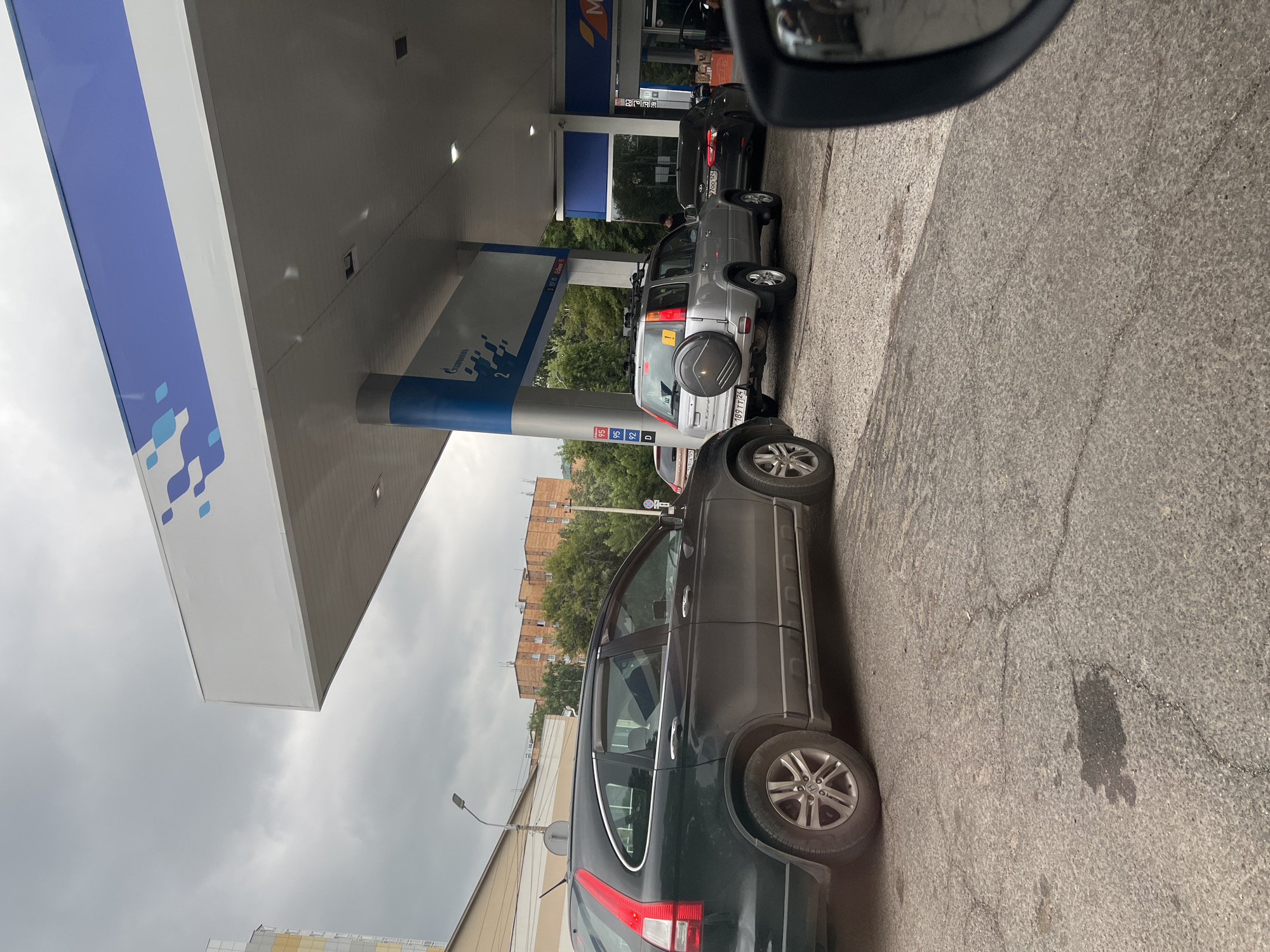 The image depicts a queue of cars at a gas station under a cloudy sky. The gas station features a white canopy with blue and white geometric patterns and a blue stripe along the top. A pillar supporting the canopy is also blue and white, with "ГАЗПРОМНЕФТЬ" (Gazpromneft) logo visible. Fuel price signs displaying "95", "92", and "D" are present.
Several vehicles are lined up: a black Honda CR-V is in the foreground, followed by a silver SUV with a spare tire mounted on the back, and a black sedan. A person is visible near the silver SUV, potentially interacting with the gas pump. Buildings and trees form the background. The ground surface is asphalt with cracks and a dark stain, possibly oil or fuel. The image appears to have been taken from inside a vehicle, as a side mirror is partially visible on the right. The presence of the Gazpromneft branding strongly suggests the location is a Gazpromneft gas station in Krasnoyarsk, Russia.