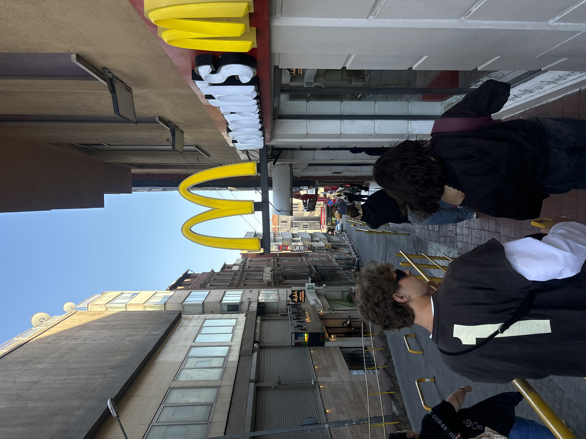 The image captures a bustling street scene in the Beyoğlu district of Istanbul, Turkey. The prominent golden arches of a McDonald's restaurant are visible, with the store's name in English on the sign. Buildings of various heights and architectural styles line the street, which slopes slightly upward into the distance. Several people are walking along the street, including a person wearing a dark shirt and a white shirt with a white number "1" on the back. The overall scene suggests a busy urban area with a mix of businesses and pedestrians.