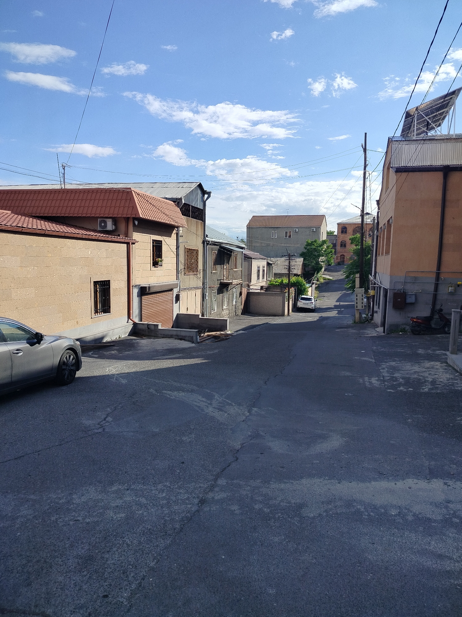The image shows a street scene in Yerevan, Armenia. The street slopes uphill and is paved with asphalt. A gray car is parked on the left side of the street in front of a single-story building with a red tile roof. Adjacent to this building is a two-story building with a roll-up door and barred windows. On the right side of the street, there is a taller building with a flat roof and solar panels. A utility pole stands near this building. A scooter is parked on the sidewalk next to the building. A white car is further up the street. In the background, multi-story buildings are visible. The sky is blue with scattered white clouds. The light suggests it is daytime.