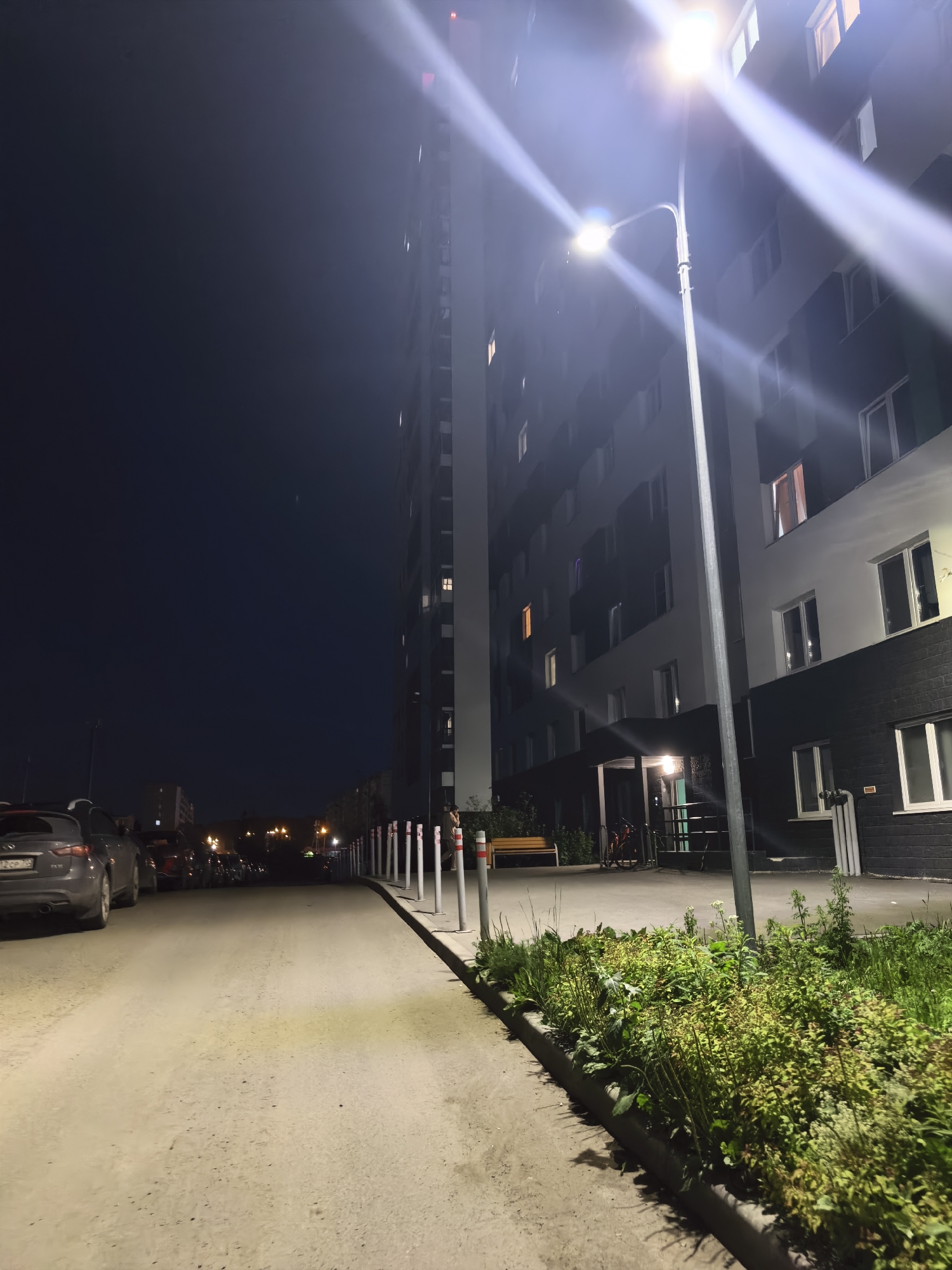 An outdoor shot at night shows a tall, dark building with white trim and numerous lit windows. A bright street lamp illuminates a paved area with parked cars and a sidewalk lined with bollards and greenery. The sky is a deep blue.