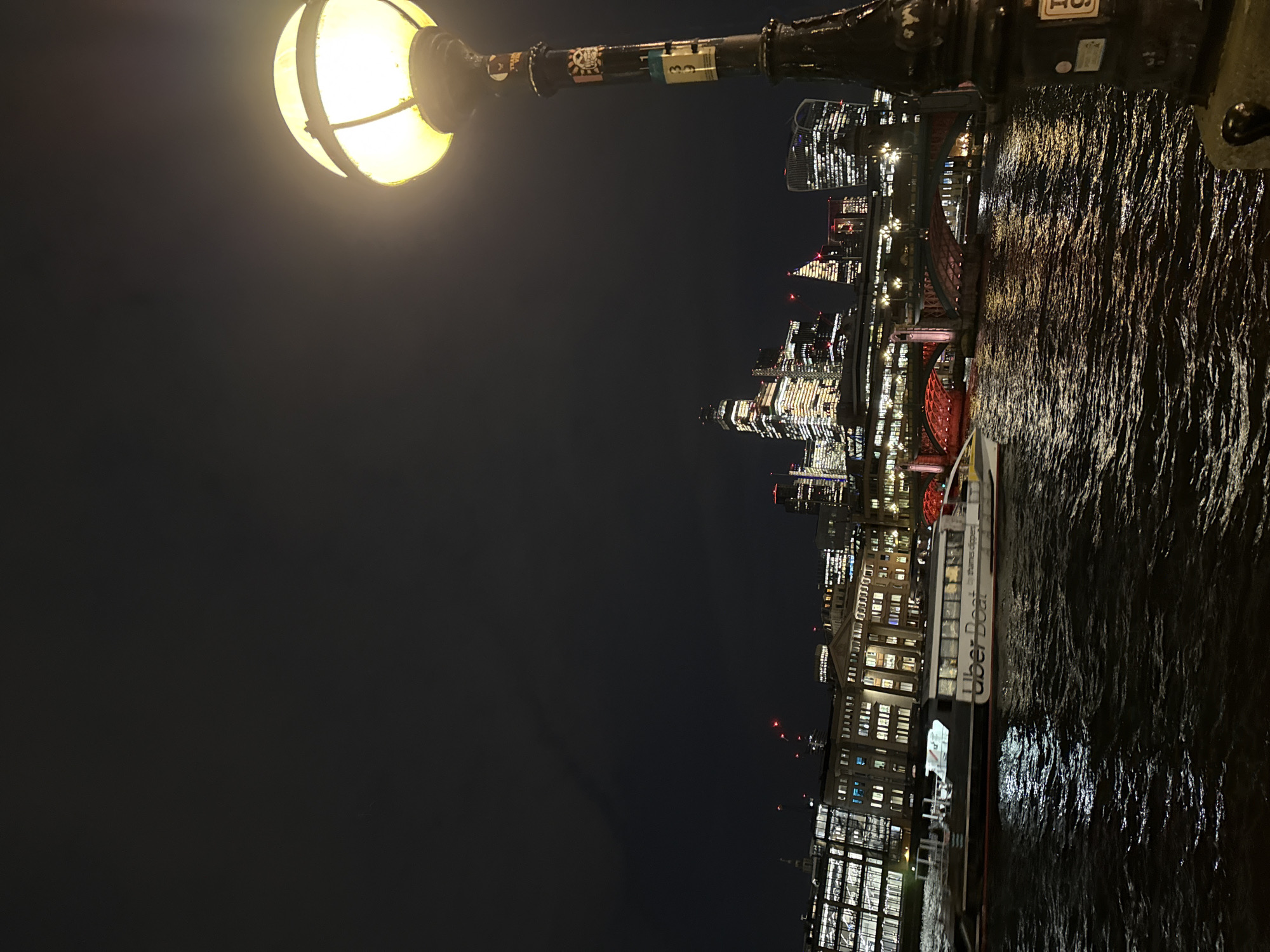 The image depicts a nighttime urban scene along the River Thames in the City of London, United Kingdom, viewed from the south bank looking towards the north bank.
In the immediate foreground, to the right, a dark, ornate streetlamp post is visible, topped with a glowing spherical amber light fixture. The lamppost itself has several stickers adhered to its surface.
The midground features the dark, rippling water of the River Thames, reflecting streaks of light from the surrounding city. A white and dark-hulled passenger vessel, clearly labeled "Uber Boat by Thames Clippers," travels from right to left across the frame. Its interior is illuminated, casting light onto the water.
In the background, the prominent illuminated skyline of the City of London is visible. Numerous skyscrapers and other commercial buildings display lit windows, contributing to a vibrant urban glow. Red warning lights are visible on the summits of several taller buildings. Directly behind the Uber Boat, a dark arched bridge, identified as Cannon Street Railway Bridge, spans the river, its underside illuminated with red lighting. Distant architectural landmarks, including the distinct shape of 20 Fenchurch Street (the "Walkie-Talkie"), are discernible within the dense collection of buildings. The sky above is dark and overcast.