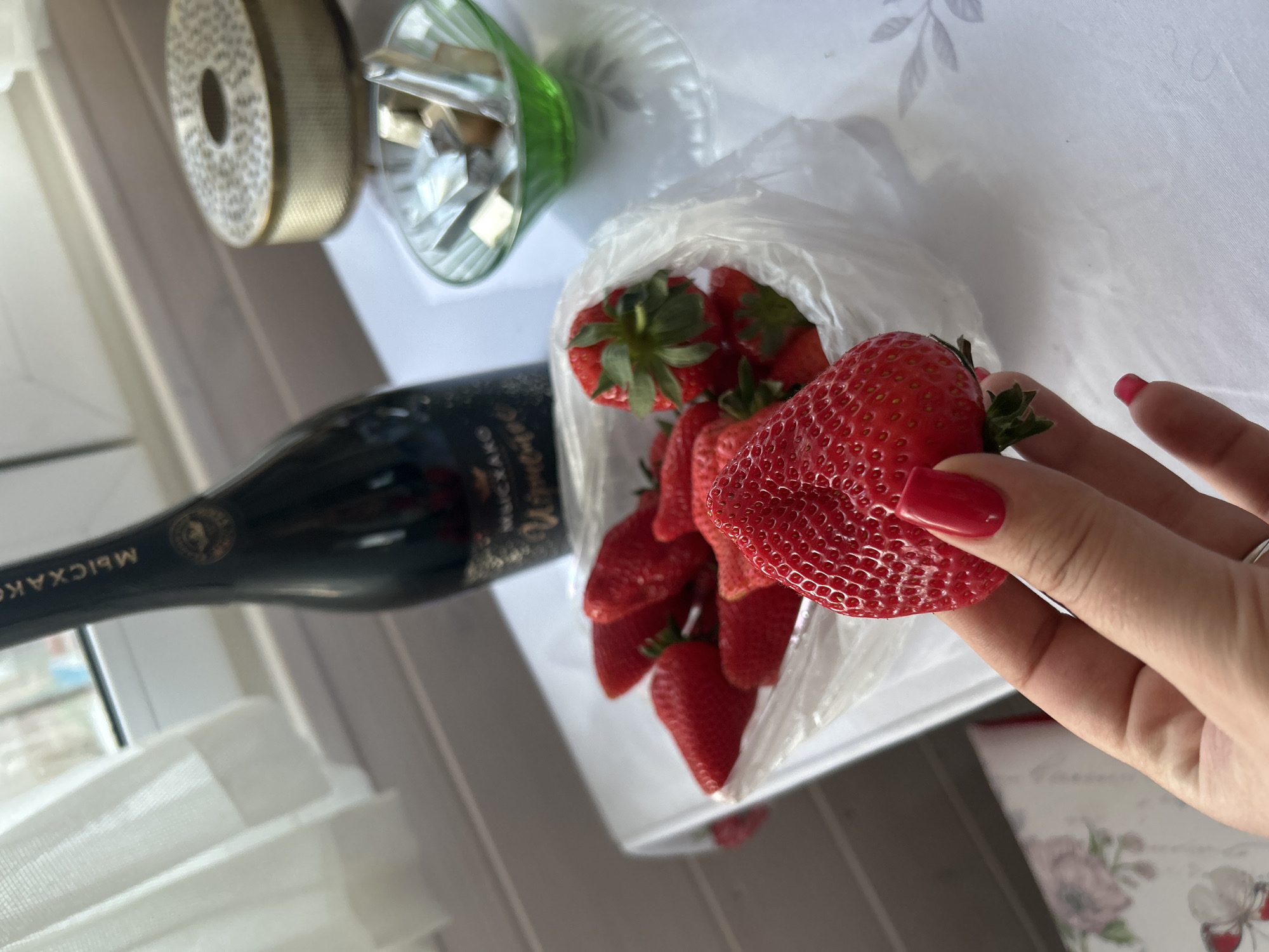 The image shows a still life arrangement on what appears to be a table indoors, potentially near a window. There is a bottle of sparkling wine, possibly champagne, with Cyrillic script visible on the label, identifying it as "Abrau-Durso," which is a Russian brand. This would indicate a possible location in Russia, though the image does not specify a location in Bashur.
Next to the bottle is a clear glass object, possibly a decorative vase. A clear plastic bag filled with fresh strawberries is centrally positioned. A hand with red nail polish holds a large strawberry. A roll of ribbon is visible in the background. The overall arrangement suggests a celebratory or festive occasion.