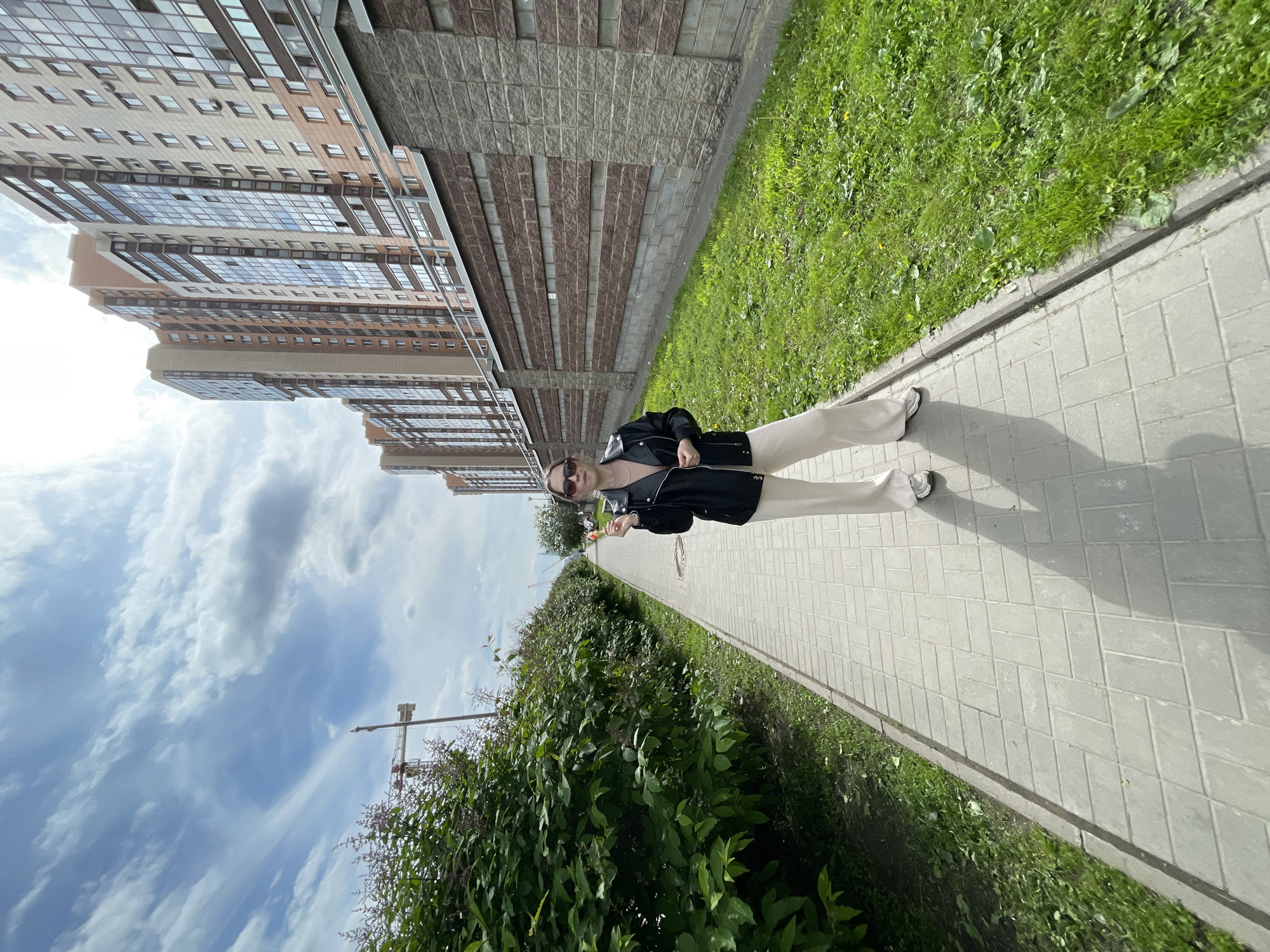 The image depicts a full-body view of a person standing on a paved sidewalk in what appears to be a modern residential area in Saint Petersburg, Russia. The scene is bright and takes place during the daytime under a partially cloudy sky.
The central subject is a young person, likely a woman, facing slightly towards the camera. She is wearing sunglasses on her head, a light-colored top (possibly beige or light pink), a black jacket, and loose-fitting, light-colored (off-white or beige) wide-leg pants. Her feet are clad in light-colored sneakers, and she holds a small, light-colored object in her right hand, possibly a phone. She is posed, standing with one foot slightly in front of the other.
The foreground features a light grey sidewalk constructed from rectangular paving blocks, stretching diagonally from the bottom towards the upper-center of the frame. To the right of the sidewalk, a narrow strip of vibrant green grass runs along a curb. Beyond the grass, a dense, leafy green hedge or row of bushes forms a natural barrier, extending into the distance.
In the background, on the left side of the image, stand several tall, modern apartment buildings. These multi-story structures exhibit facades composed of light-colored panels and sections of reddish-brown or dark-grey brickwork, punctuated by numerous windows that reflect the sky. Above the buildings and bushes, the sky is a pale blue with scattered white, fluffy clouds. Faintly visible through the bushes against the sky is a metallic structure, possibly part of a construction crane or a utility pole.
Distinct shadows are cast on the sidewalk and grass, indicating bright daylight, suggesting the time of day is either late morning or afternoon. The overall setting is urban and contemporary. No visible text is present in the image.