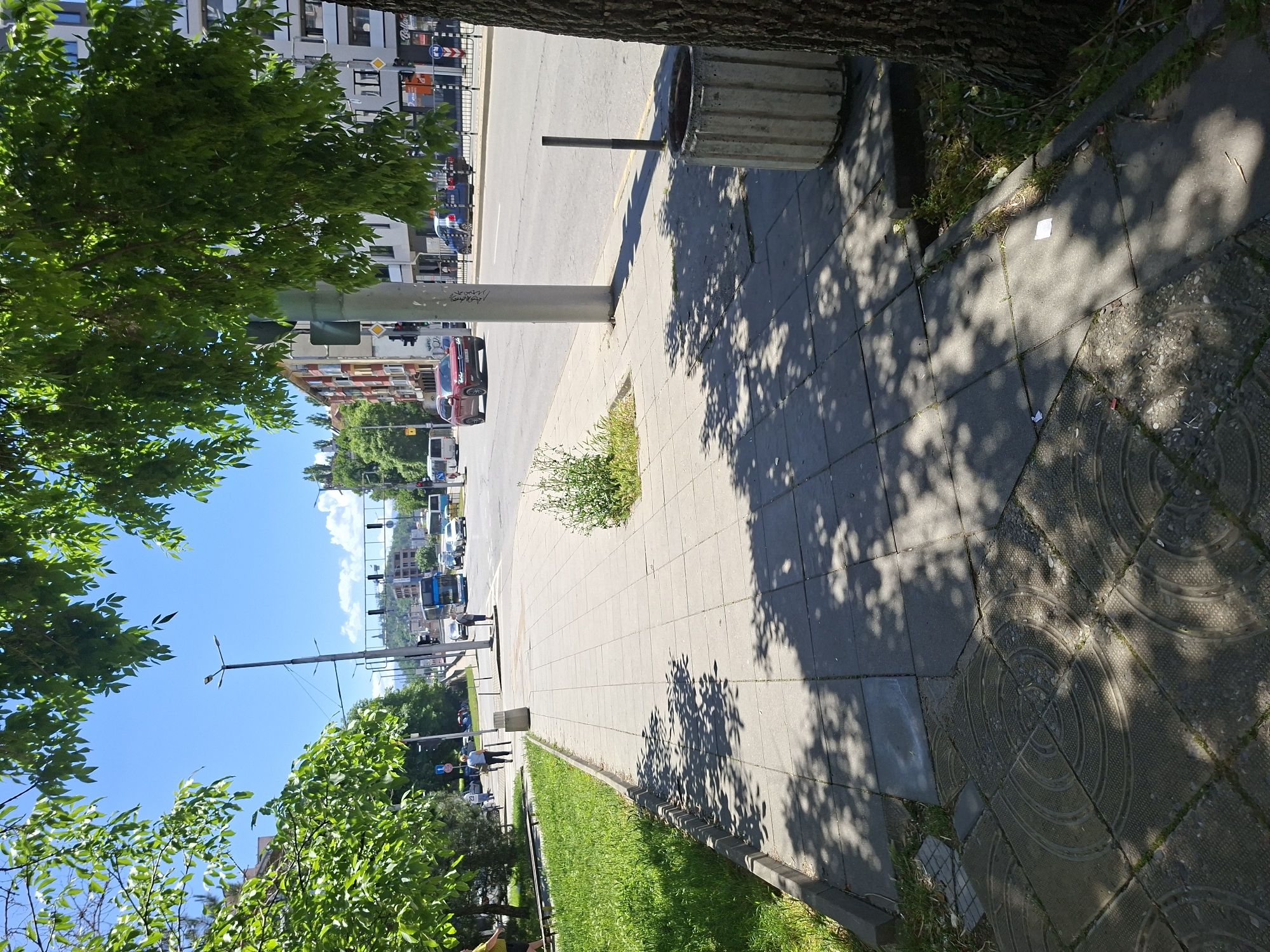 The image captures a street scene in Plovdiv, Bulgaria. The photo appears to be taken from a high angle, looking down a pedestrian sidewalk. The sidewalk is constructed with grey paving stones and is partially shaded by tree foliage. Adjacent to the sidewalk is a grassy area and a tree trunk, further contributing to the shadows cast across the pavement. In the middle ground, a concrete trash bin stands near a vertical metal pole. Beyond the sidewalk, a street with traffic is visible, featuring cars, vans, and possibly buses. Buildings of varying heights and architectural styles are seen across the street. The sky is blue with some clouds. The scene suggests a typical day in an urban environment in Plovdiv.