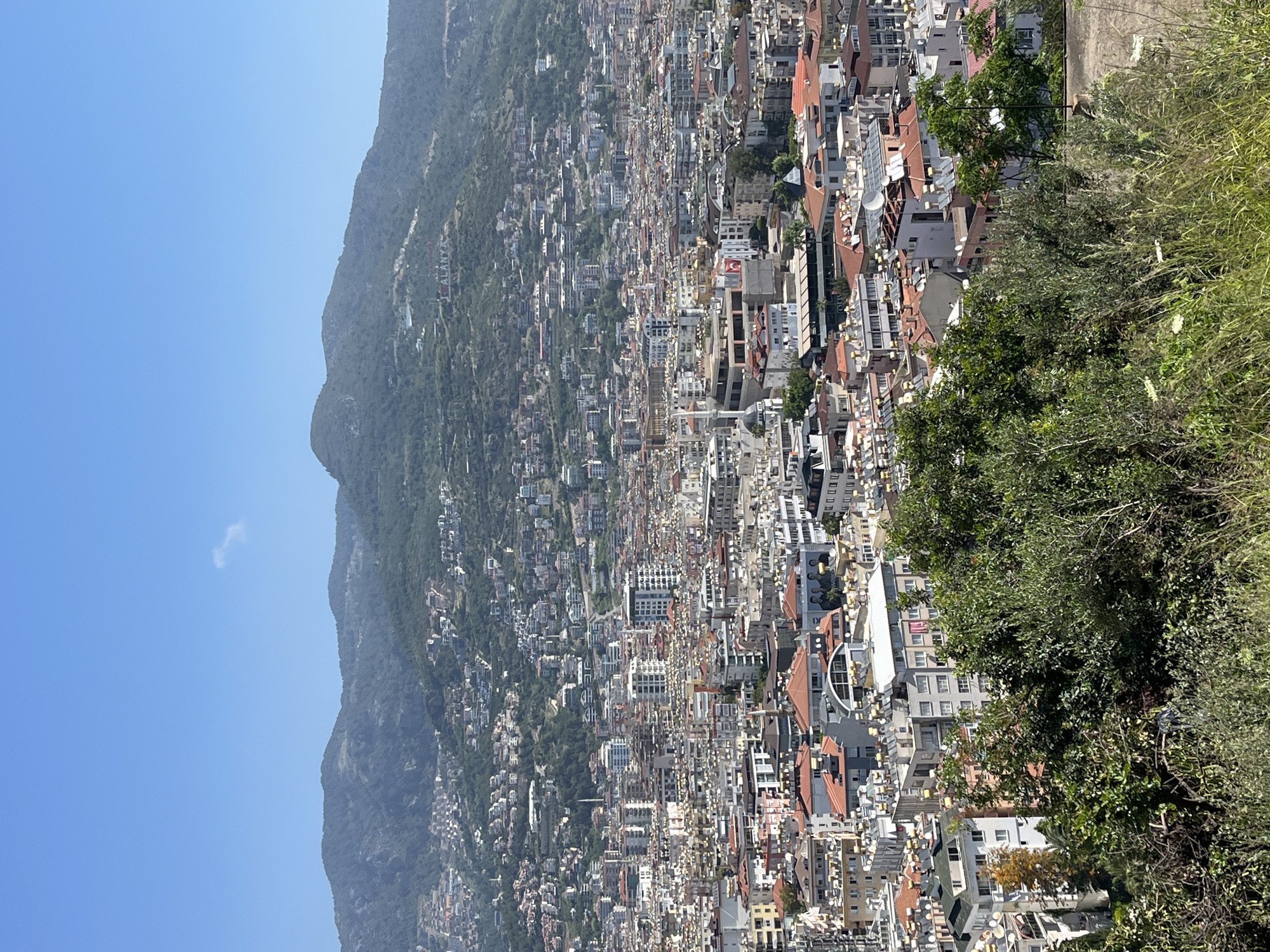 The image shows an aerial view of Alanya, Turkey, from a high vantage point, likely a mountain or elevated location. The landscape features a dense urban sprawl of buildings, mainly white and light-colored structures, densely packed. Red-tiled roofs are scattered throughout the city. A mountain covered with buildings and vegetation rises in the background. The right side of the image contains greenery, suggesting the presence of trees and bushes close to the viewing point. The sky is clear and blue. On the mountain, there is a white sign that reads "I ❤️ ALANYA".