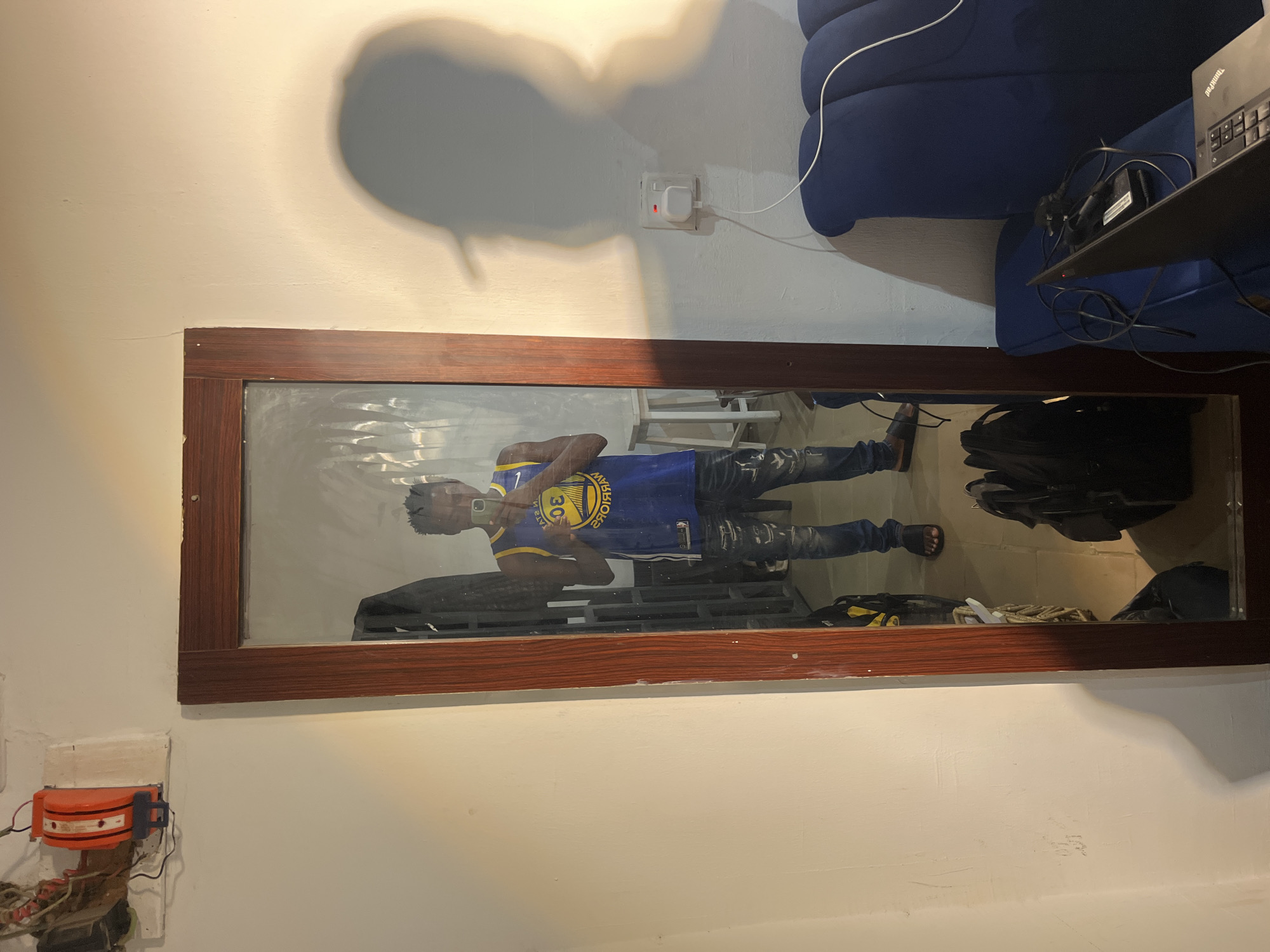 This media file is a full-body mirror selfie of a young Black man. He is standing and taking the photo himself, so his reflection is the primary subject. He is wearing a blue and yellow basketball jersey with the number "30" and the name "Curry" on the back, along with distressed denim jeans and black sandals. He has short black hair.
The setting appears to be indoors, in a room with white walls. In the background of the mirror's reflection, there is a dark blue couch and a laptop on a surface. There are also electrical outlets and some wires visible on the wall. The lighting suggests it could be daytime, possibly with artificial light contributing to the illumination. The overall mood is casual and everyday.
The main activity is the man taking a selfie. There is no discernible text other than the name and number on his basketball jersey, which is "Curry" and "30" respectively, indicating he is wearing a Stephen Curry jersey.