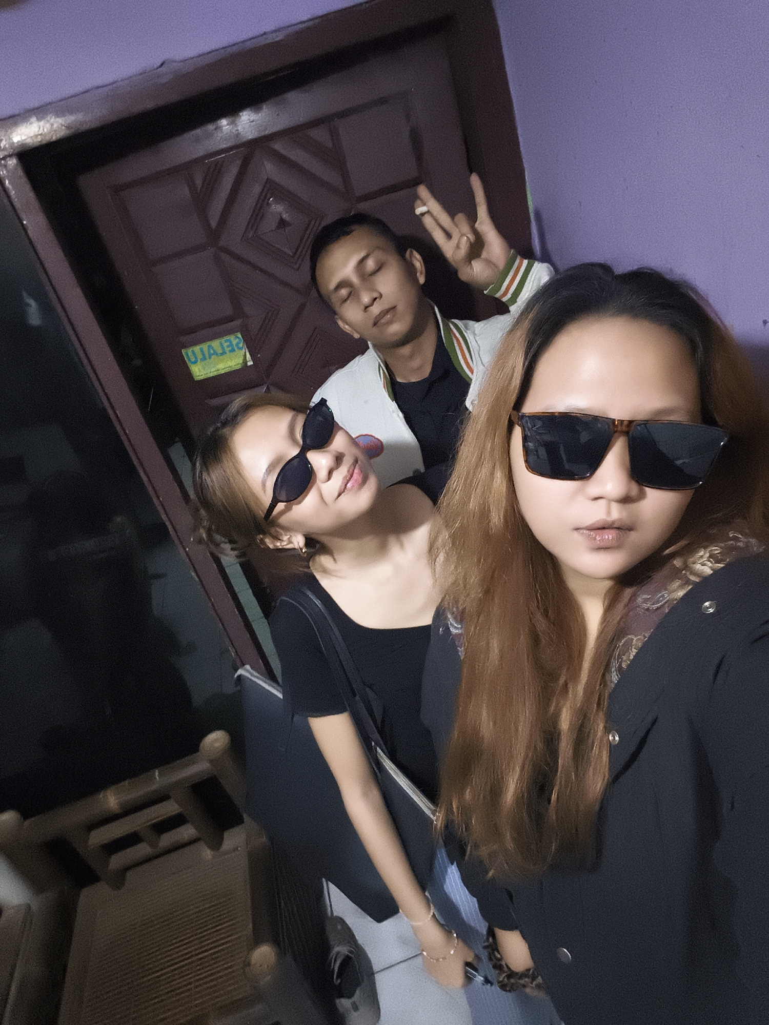 Three individuals are captured in a dimly lit interior, possibly a residence, with a purple-painted wall and a dark wooden door dominating the background. The door has a geometric pattern carved into it and a green sticker with reversed lettering that reads "UJAJE". A man is positioned behind and slightly above the other two, with his eyes closed and holding up two fingers in a peace sign, a cigarette visible between them. He is wearing a jacket with green and red striped trim. In the foreground, two women are taking a selfie. The woman on the left, with light brown hair pulled back, wears black sunglasses and a black top, holding a bag. The woman on the right, with long, light brown hair and wearing large, dark sunglasses, is also in a black top, and appears to be wearing a patterned scarf or jacket. The overall impression is casual, with the subjects possibly posing for a photograph. To the left of the frame, a portion of what appears to be a wooden chair is visible.