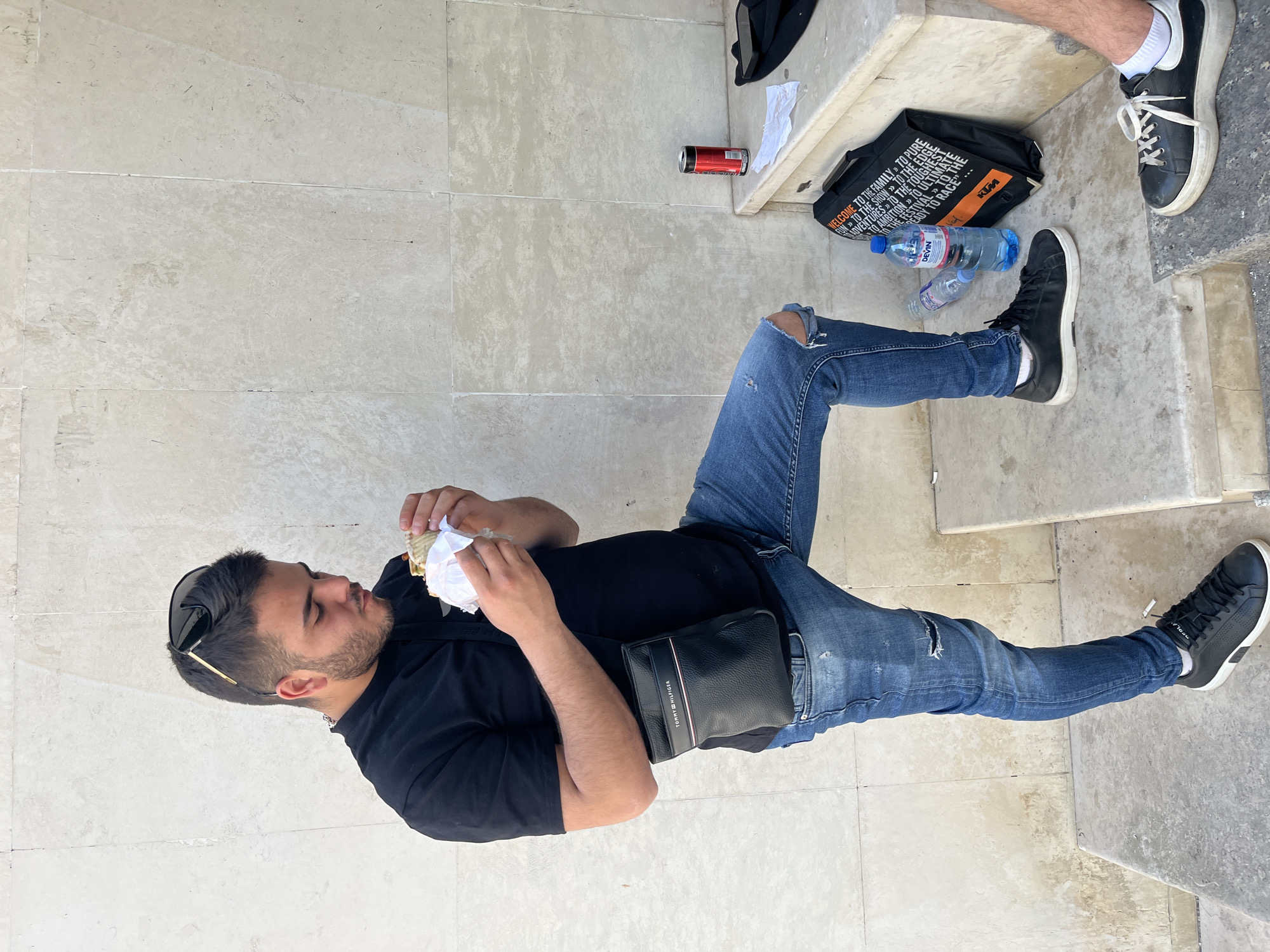 A man sits on a stone ledge in Plovdiv, Bulgaria, eating a sandwich. He wears ripped jeans, a black shirt, and black sneakers. A black Tommy Hilfiger bag rests on his lap. Nearby are water bottles, a KTM bag, and a can. Another person's legs are visible next to the bag. The setting suggests an urban, possibly touristy area due to the stone construction.