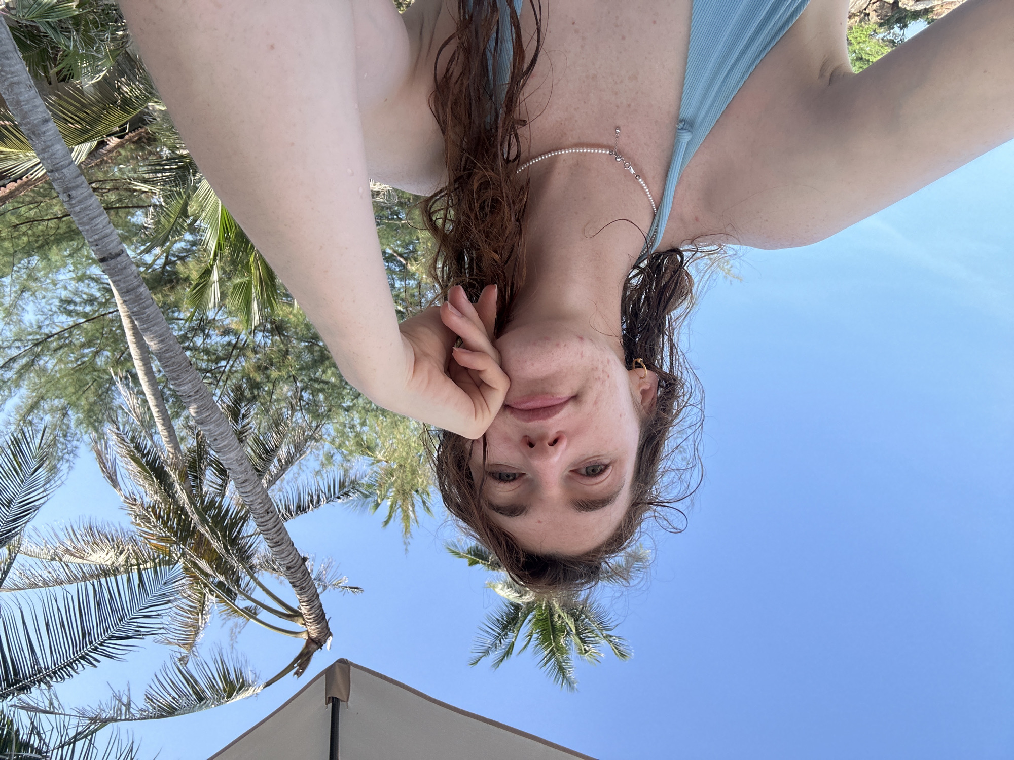 A low-angle, upside-down photograph shows a woman at Baan Chaweng Noi, Thailand. She is wearing a light blue swimsuit and a pearl necklace, with wet, reddish-brown hair. The woman's face is visible, and she has her hand near her face. The background features a bright blue sky, palm trees, and a portion of a beige sun umbrella.