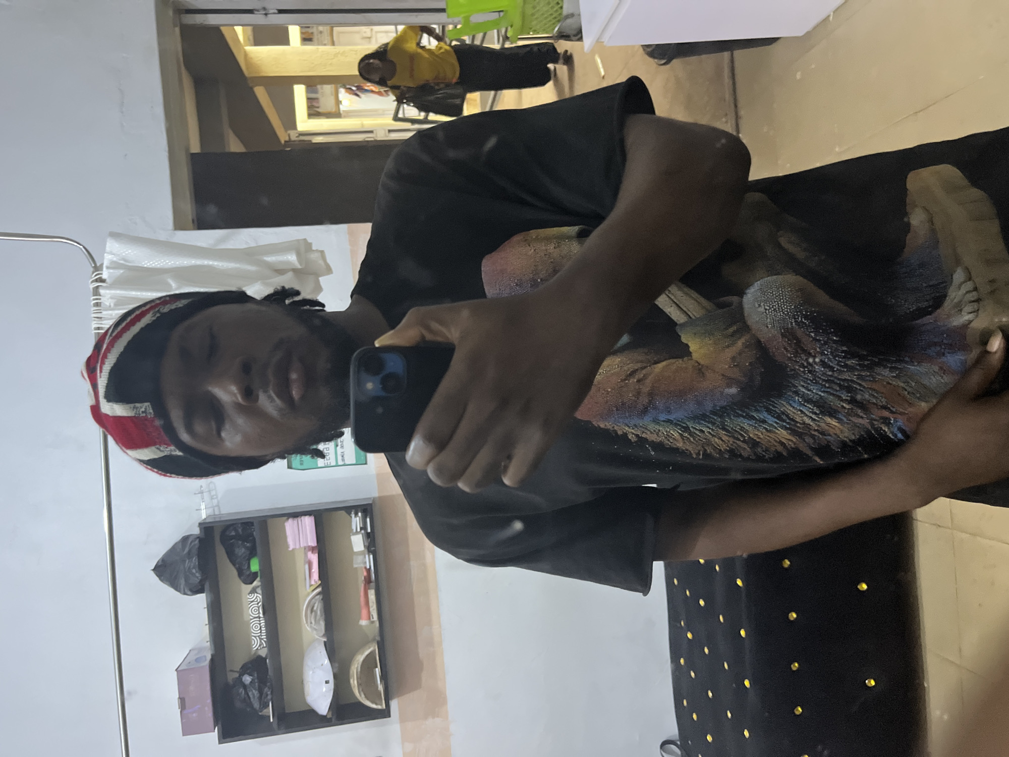 In the city of Jos, Nigeria, a young Black man is captured in a selfie taken in what appears to be a room with a shelf unit. The man is wearing a black t-shirt with a large graphic print and a multi-colored beanie. He holds a smartphone in his right hand, using it to take the photograph. Behind him, a shelf unit displays various items, including what seem to be packaged goods and a black trash bag. To the right, a doorway reveals a hallway and a woman in a yellow shirt and dark pants walking away, carrying a dark bag. The overall environment suggests a casual, indoor setting, possibly a residential or shop area within Jos.