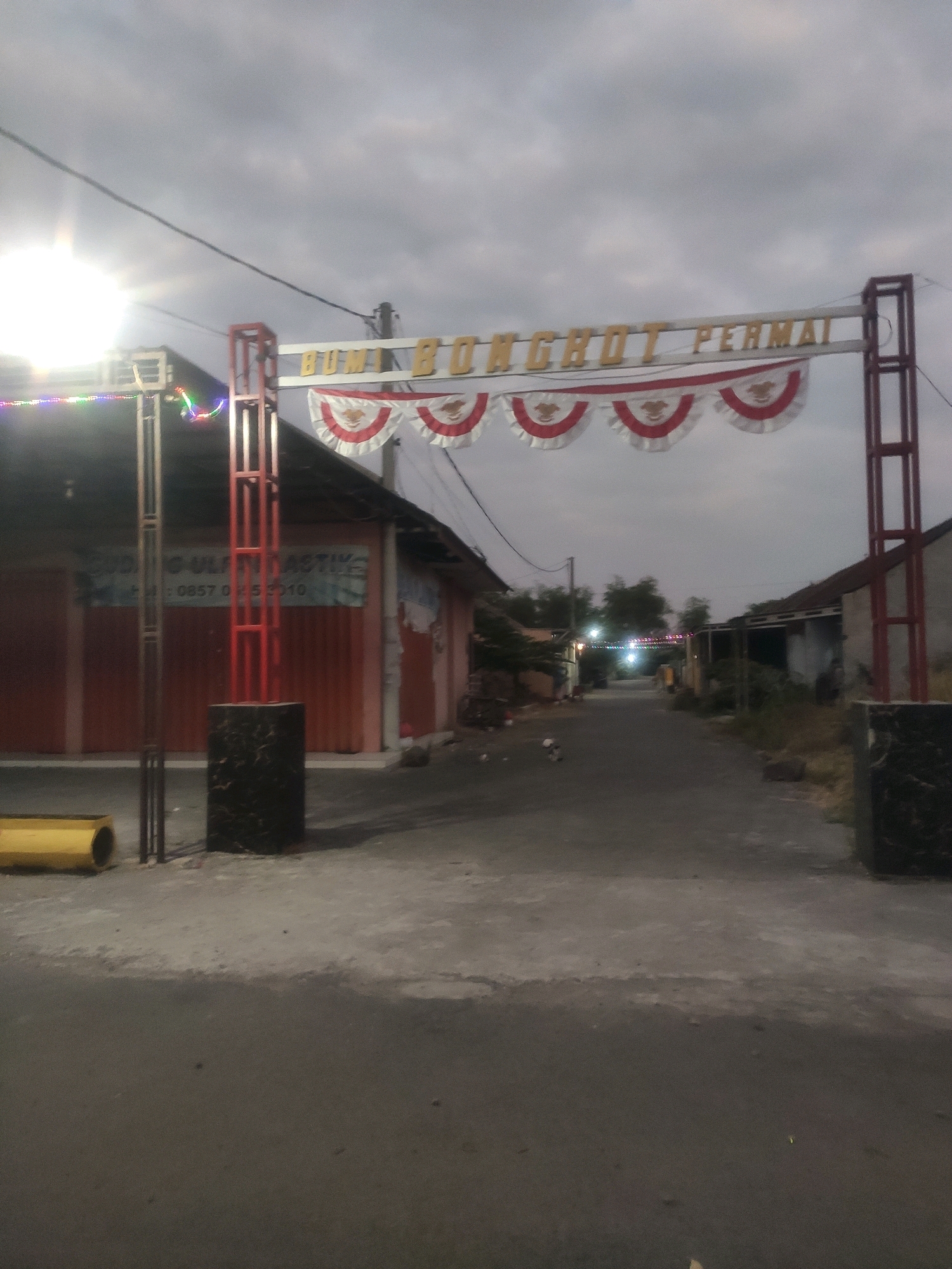 An outdoor, eye-level, medium shot captures an archway erected over a street at dusk. The archway, constructed of red metal framing, supports a horizontal beam with the words "BUMBON" and "PERMAI" in gold lettering. A string of red and white pennant banners hangs from the beam. On the left side of the arch, a building with a red facade is visible, featuring a blue banner with text that is partially obscured but includes a phone number. A bright light source is visible to the left of the arch. The street leads into the distance, lined with buildings and trees, and illuminated by decorative lights. The sky above is overcast and gray.