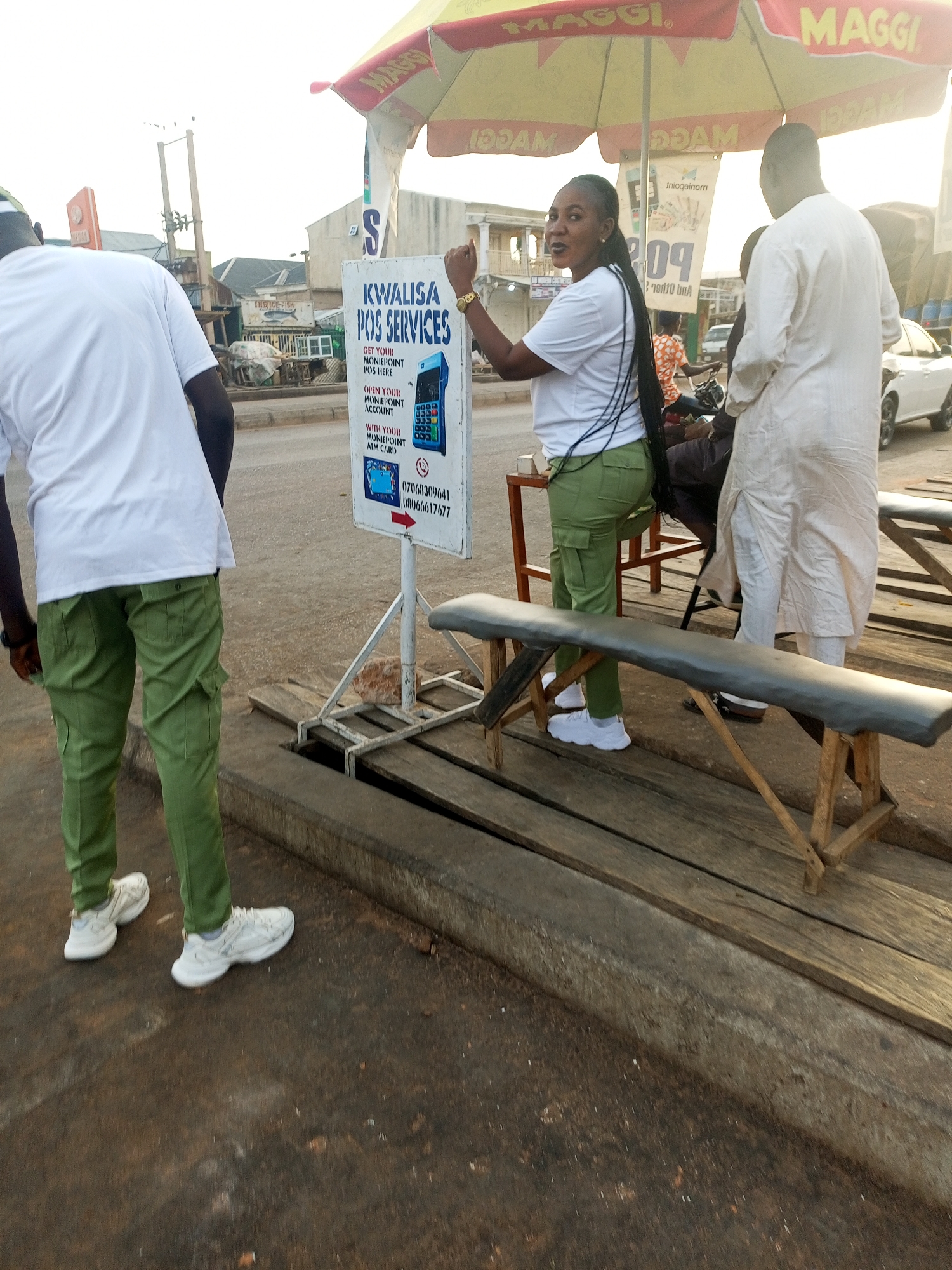 The image shows a street scene in Gombe, Nigeria, where a woman in a white t-shirt and green trousers is operating a Kwelisa POS service under a "Maggi" branded umbrella. Next to her is a man in white attire. A person wearing the same green trousers and white shirt is entering the scene. There is a sign advertising "Kwalisa POS Services" with phone numbers displayed. Wooden benches are placed nearby for customers. The scene is set on a raised wooden platform adjacent to the street. Other buildings and a vehicle are visible in the background.