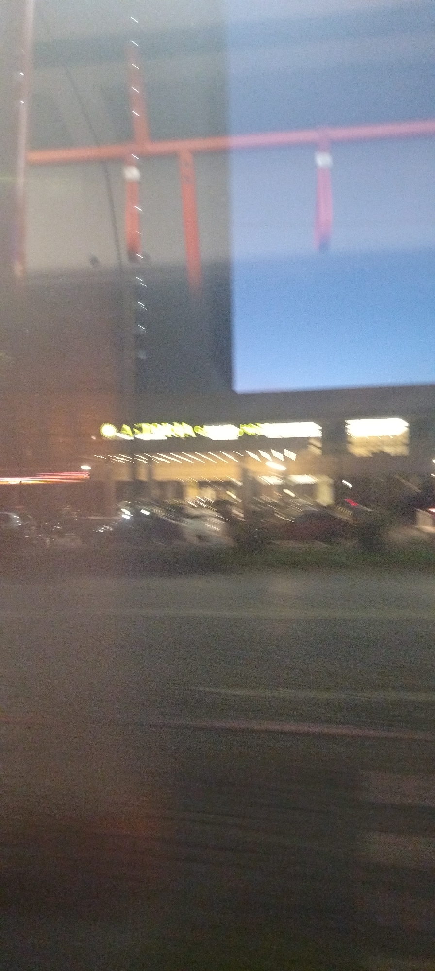 The image shows a blurred nighttime view of a street in front of a building with illuminated signage. The signage appears to read "ASTOR G". There are cars parked in front of the building, and the street in the foreground is dark and appears to be asphalt. The background includes an orange structure with vertical white lights. The overall impression is one of a commercial area at night, possibly a hotel or entertainment venue.