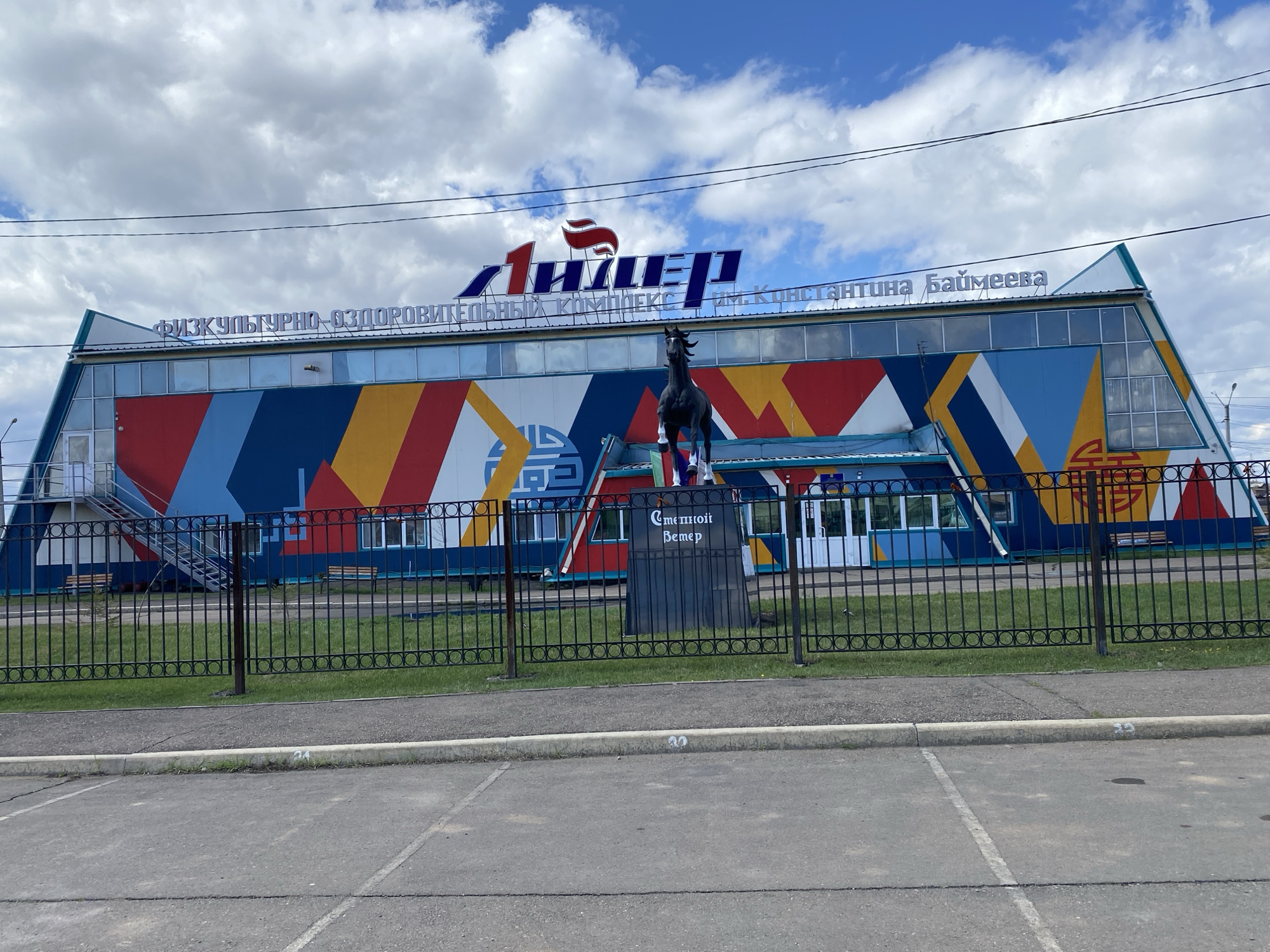 The image depicts the "Лидер" (Leader) Sports and Recreation Complex named after K. Baymeev, located in Ust-Ordynsky, Russia. The building features a colorful facade of blue, red, yellow, and white geometric shapes. Above the entrance, signage indicates the complex's name in Russian. A black horse statue stands behind a black metal fence in front of the building, with a plaque reading "Степной ветер" (Steppe Wind). The ground in front of the fence is paved with grey concrete, and some greenery is visible behind the fence. The sky above is partly cloudy.