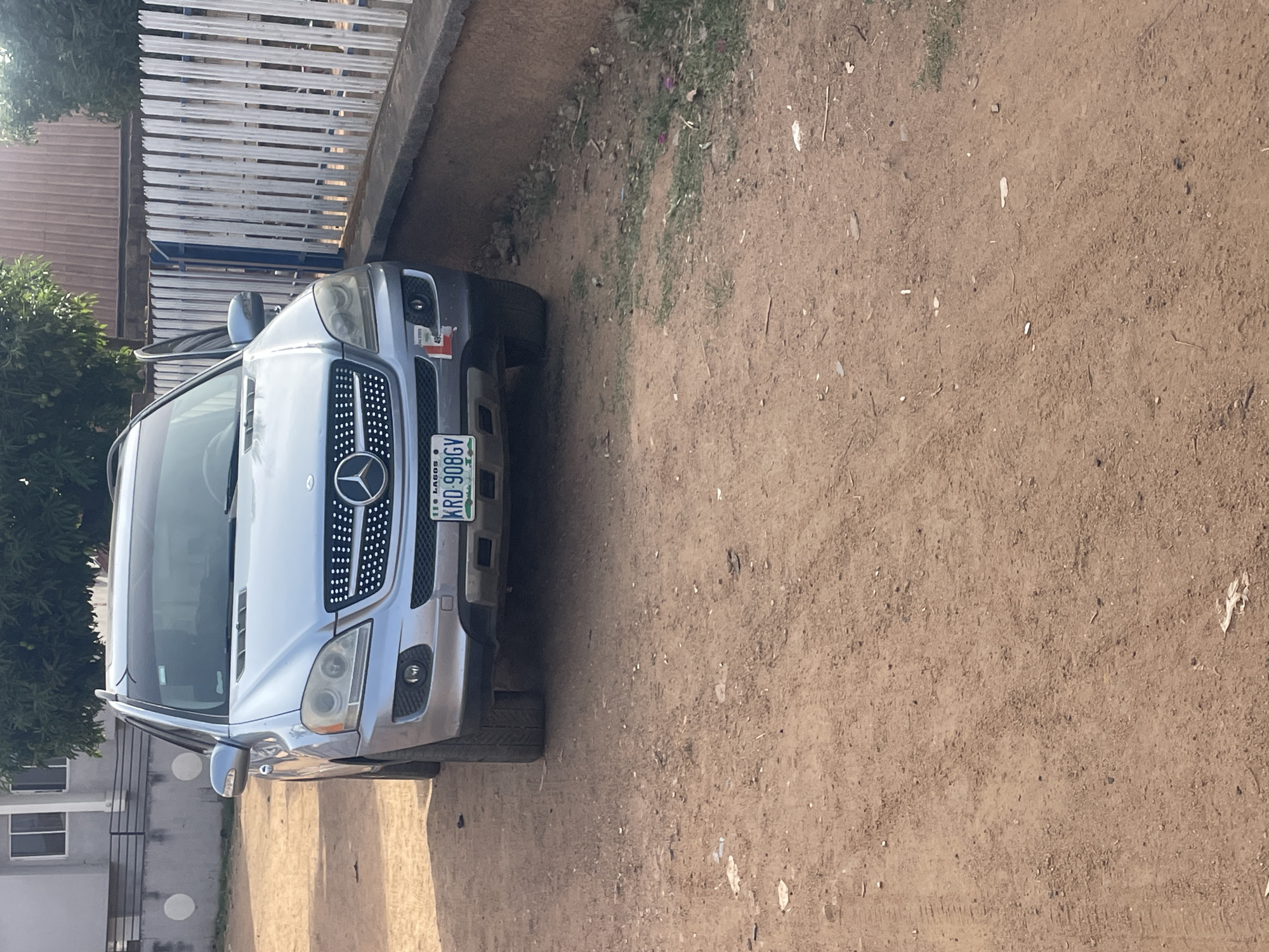 An outdoor image, rotated 90 degrees counter-clockwise, depicts a light-colored Mercedes-Benz SUV parked on an unpaved, reddish-brown dirt surface. The vehicle, appearing to be silver or light blue, faces slightly towards the right, showcasing its front grille with the prominent Mercedes-Benz star logo, headlights, and front bumper.
The front license plate clearly displays the alphanumeric code "KRD-908GY" with "LAGOS" written beneath it.
In the background, a wooden slatted fence runs across the upper part of the frame, behind the vehicle. To the left of the car, lush green foliage from trees or bushes is visible. A building with white-framed windows is partially seen in the lower-left corner, suggesting a residential or other structure. The ground is mostly bare earth with some scattered debris and faint tire tracks, along with a few sparse patches of green grass. The scene is illuminated by bright daylight, indicating clear, sunny weather. The setting appears to be a semi-urban or residential area in Jos, Nigeria, though the vehicle's registration is in Lagos.