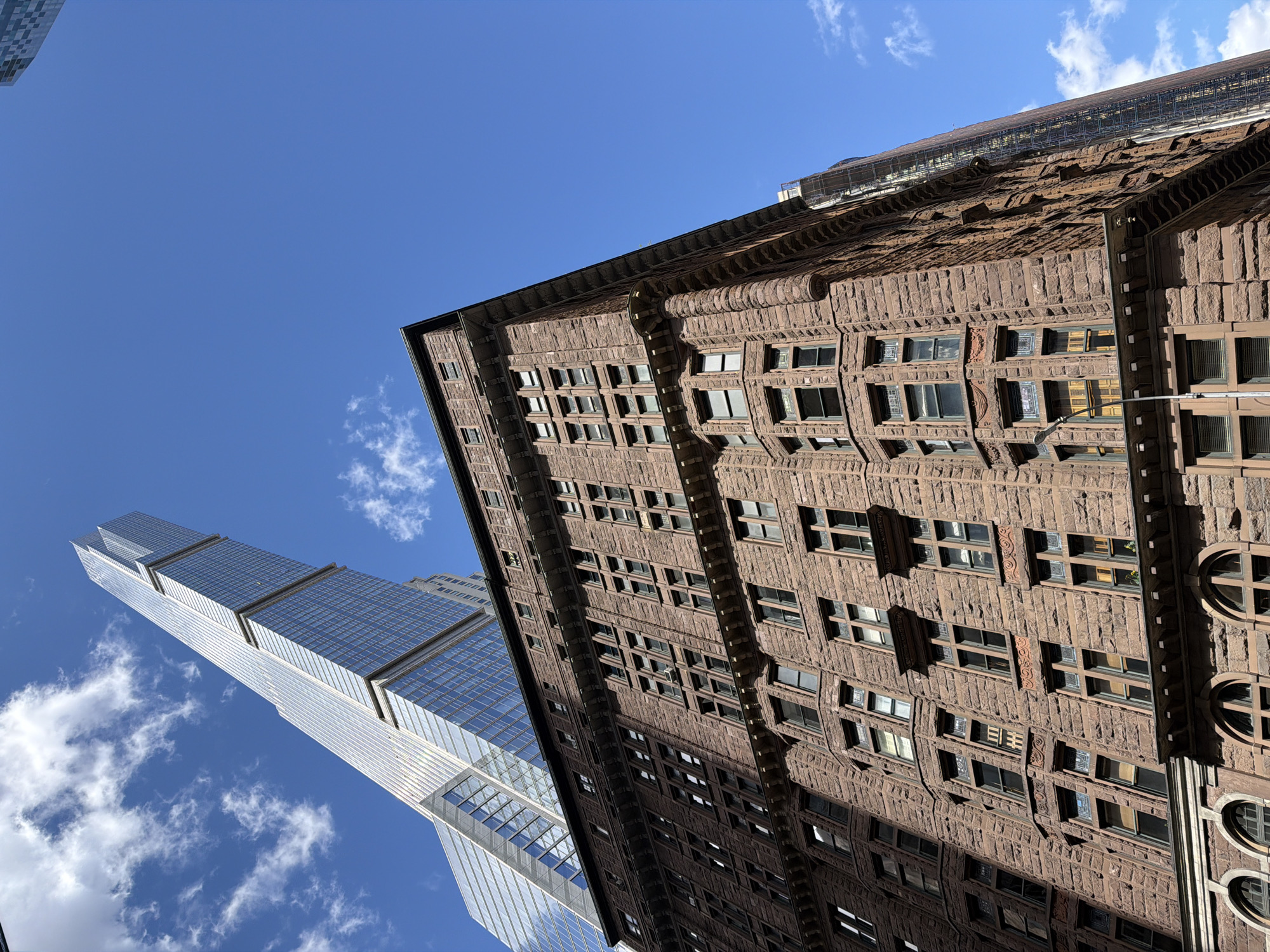 The image captures a low-angle view of buildings in New York City against a blue sky. The building on the right is older, featuring a brown stone facade with numerous windows and intricate architectural details. To the left, a modern skyscraper rises into the sky, its glass exterior reflecting the blue sky. The sky is mostly clear with a few scattered clouds.