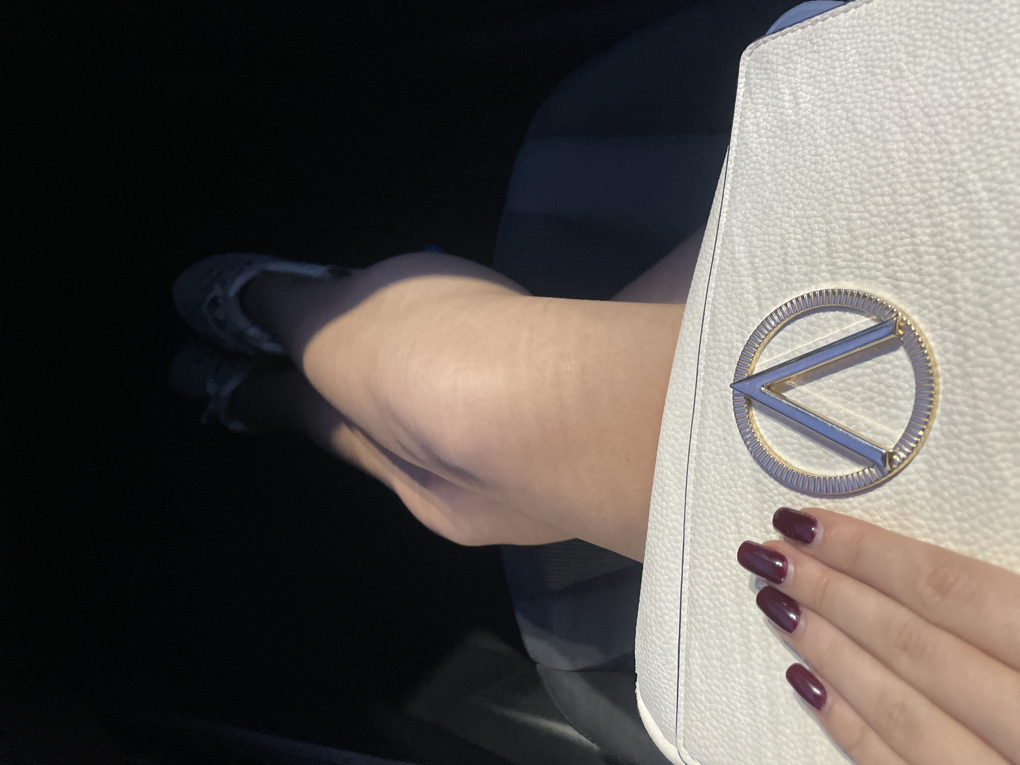This close-up image captures a person's legs and hand inside what appears to be a vehicle, likely a car, on a sunny day in Sofia, Bulgaria. Bright sunlight illuminates the scene, casting distinct highlights and shadows.
The subject's legs are extended and partially visible, covered in dark, possibly opaque, hosiery or leggings. The skin on the exposed parts of the legs is fair. Dark, patterned shoes or flats are glimpsed on the feet. The legs rest on what seems to be the dark fabric of a car seat.
In the foreground, a hand with long, neatly manicured nails painted in a deep burgundy or plum color holds a white handbag. The handbag has a textured, pebbled surface and features a prominent metallic logo. The logo consists of a gold-toned circular frame with a ridged edge, enclosing a sleek, silver-toned 'V' or chevron-shaped emblem.
The background is dark and out of focus, indicating the limited space of a car interior. No specific texts or additional location cues are visible within the image itself. The overall activity depicted is a person sitting as a passenger, with their belongings, possibly during travel or a commute.